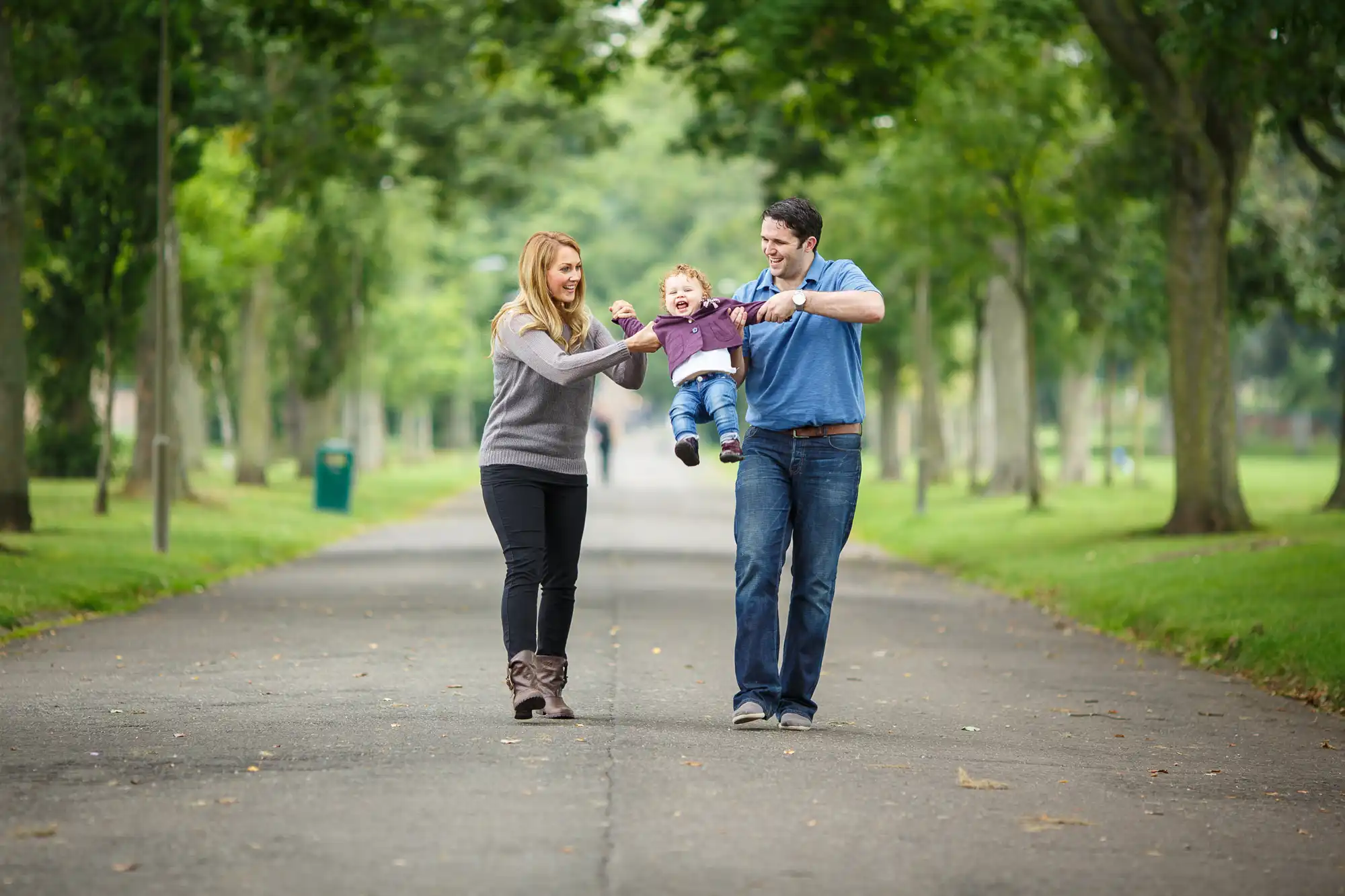 A young couple swinging their toddler in the air while walking on a tree-lined path in a park.