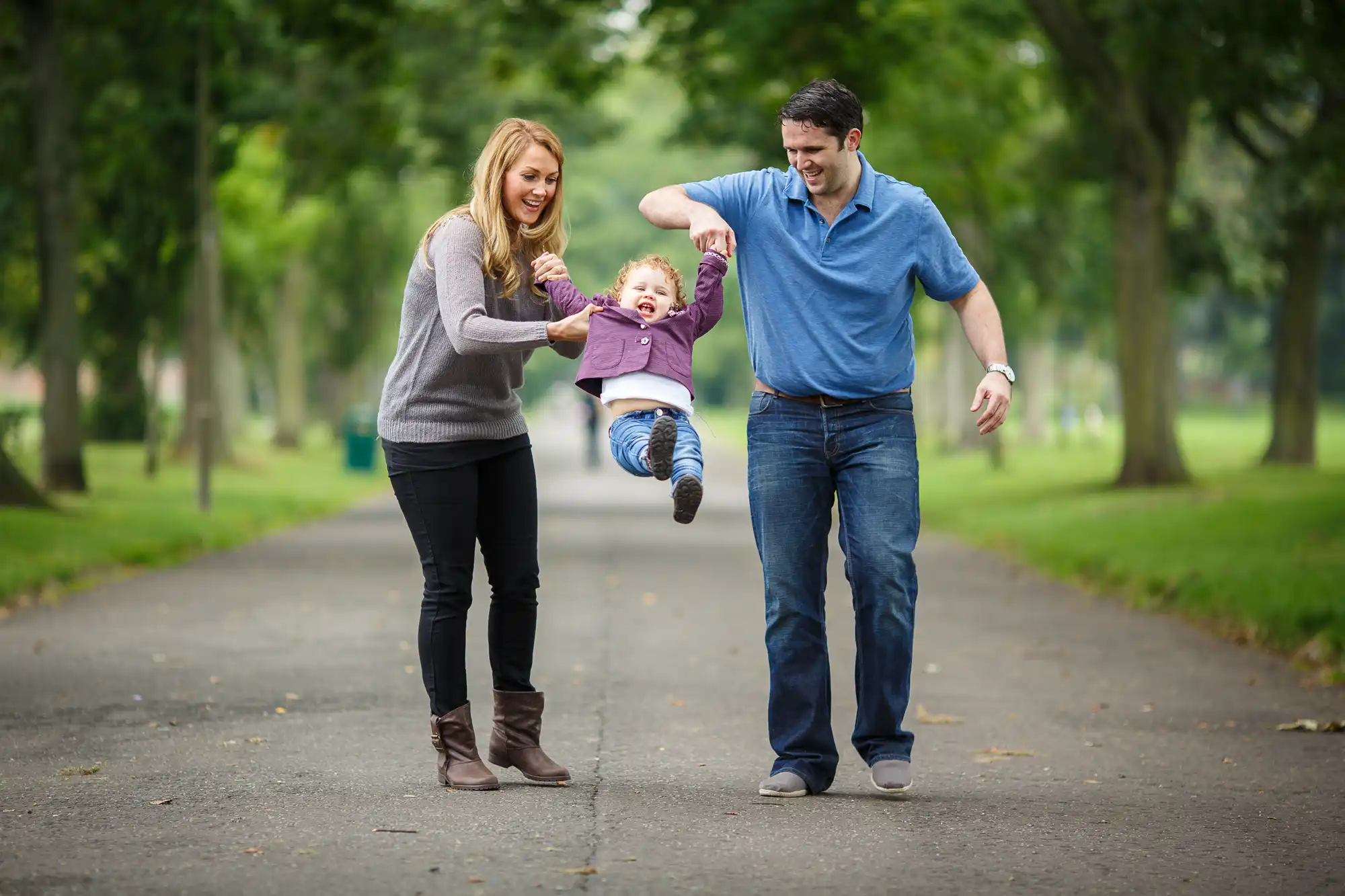 A joyful family swings their young child by the arms while walking on a park pathway, surrounded by trees.