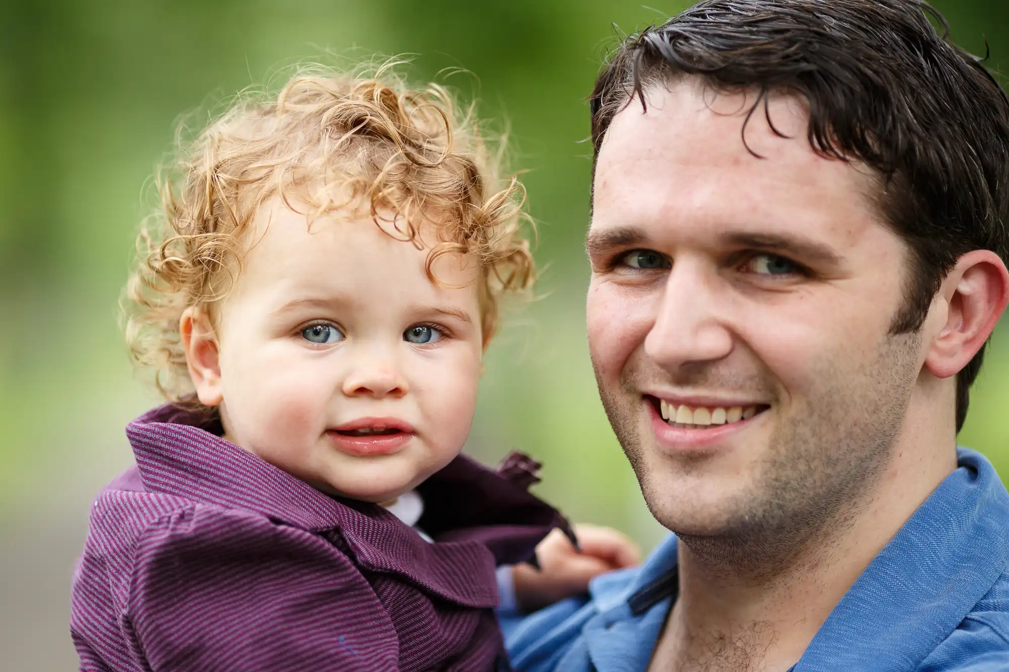A father holding his toddler with curly hair, both smiling slightly, outdoors with a blurred green background.