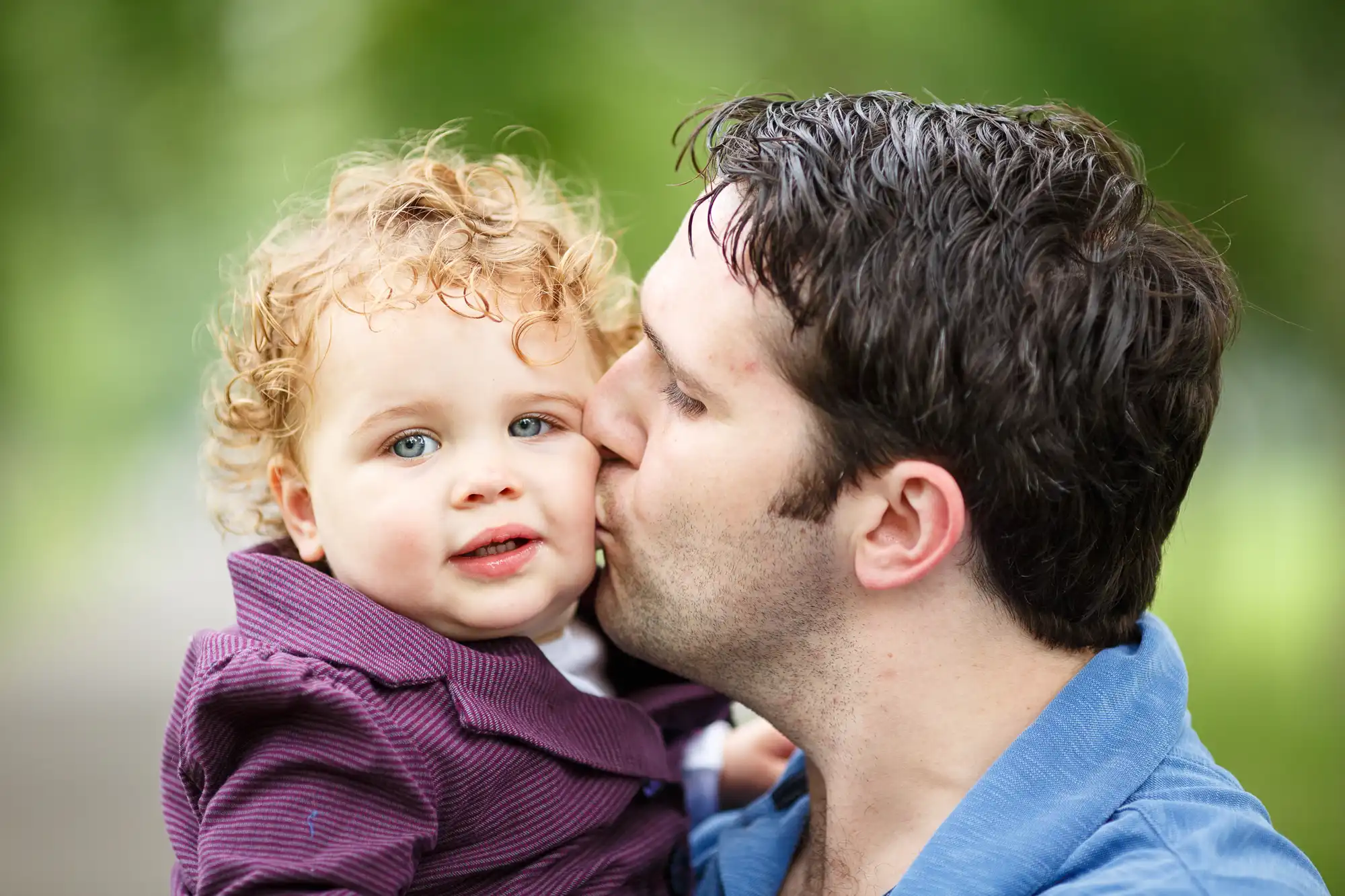 Father kissing his toddler with curly hair on the cheek, both smiling, outdoors with blurry green background.