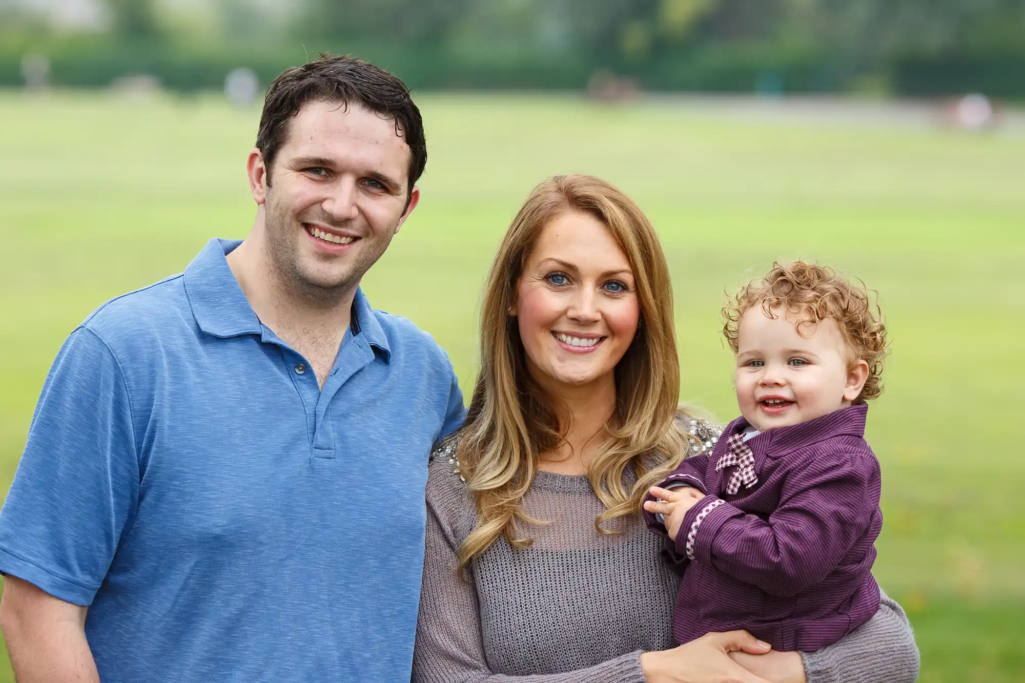 A family of three smiling outdoors; a man in a blue polo, a woman with blonde hair in gray, and a toddler with curly hair in purple.