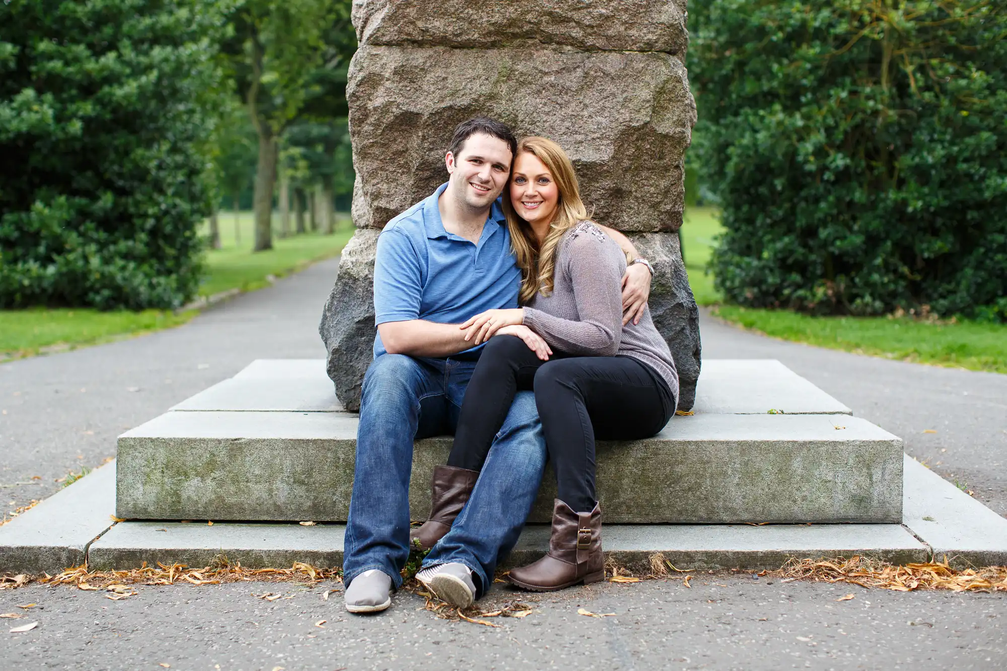 A couple smiling and sitting together on a concrete step by a stone column in a park, surrounded by trees.