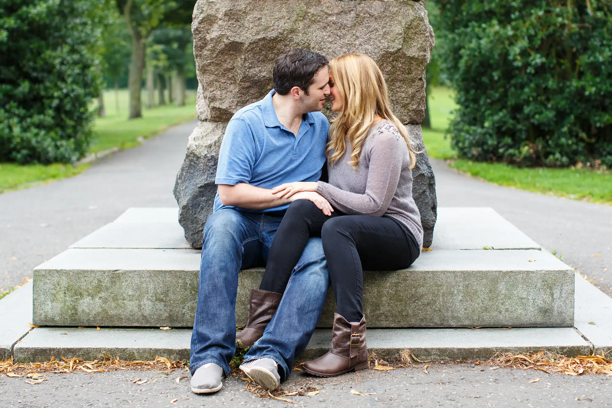 A couple sitting close and kissing on a bench in a park with a large stone structure behind them and trees along the pathway.