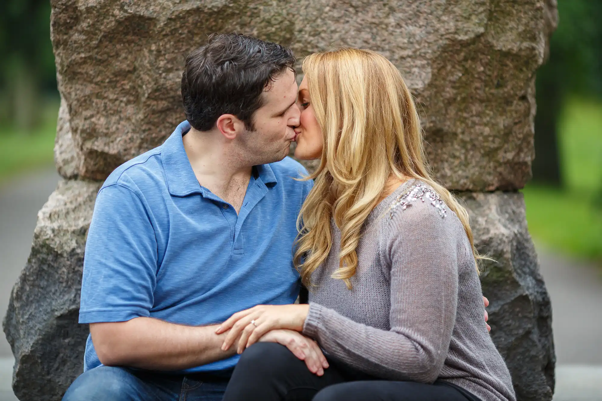 A couple kissing while seated on a bench in a park, with a large rock in the background. they are both wearing casual shirts.