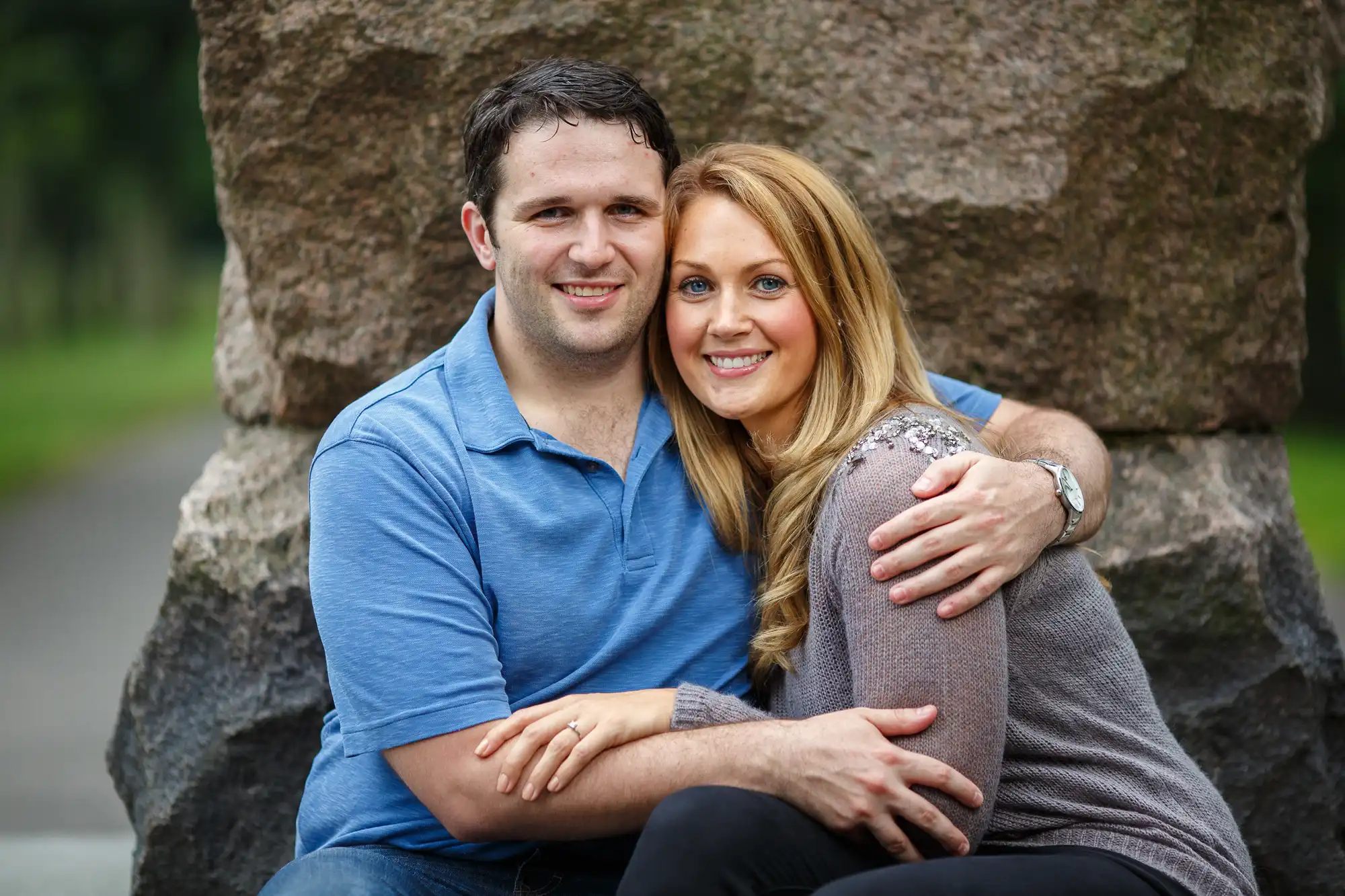 A man and a woman smiling and embracing while sitting on a large rock in a park.
