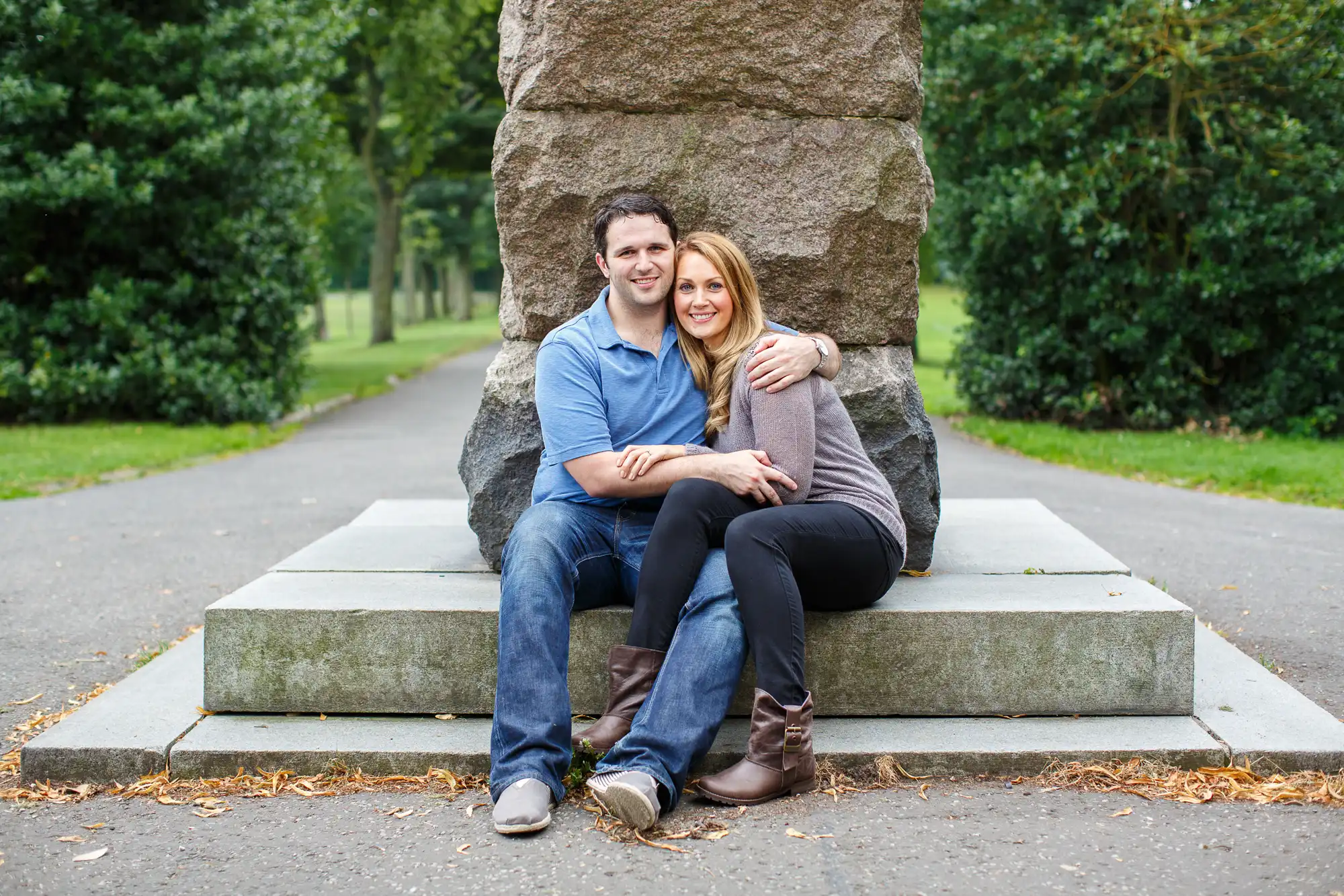 A couple sitting closely on the steps of a stone monument in a park, smiling at the camera.