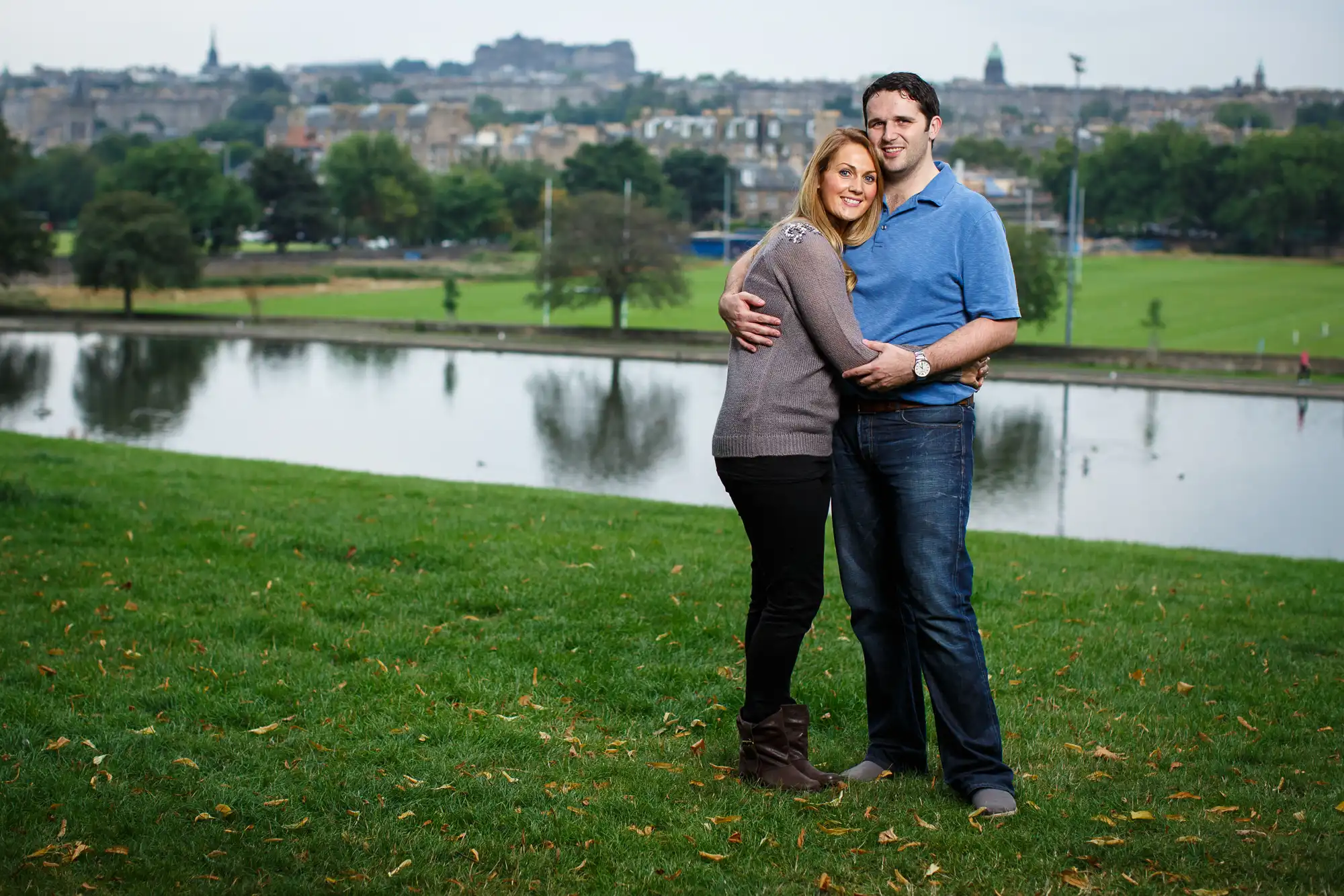 A couple embracing outdoors on a grassy hill, overlooking a cityscape and river in the background.