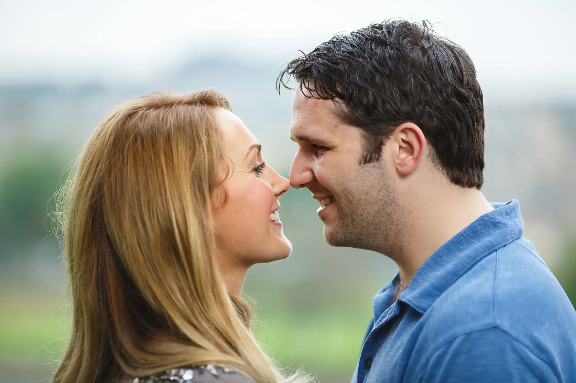 A man and a woman smiling closely at each other outdoors with a softly blurred background.