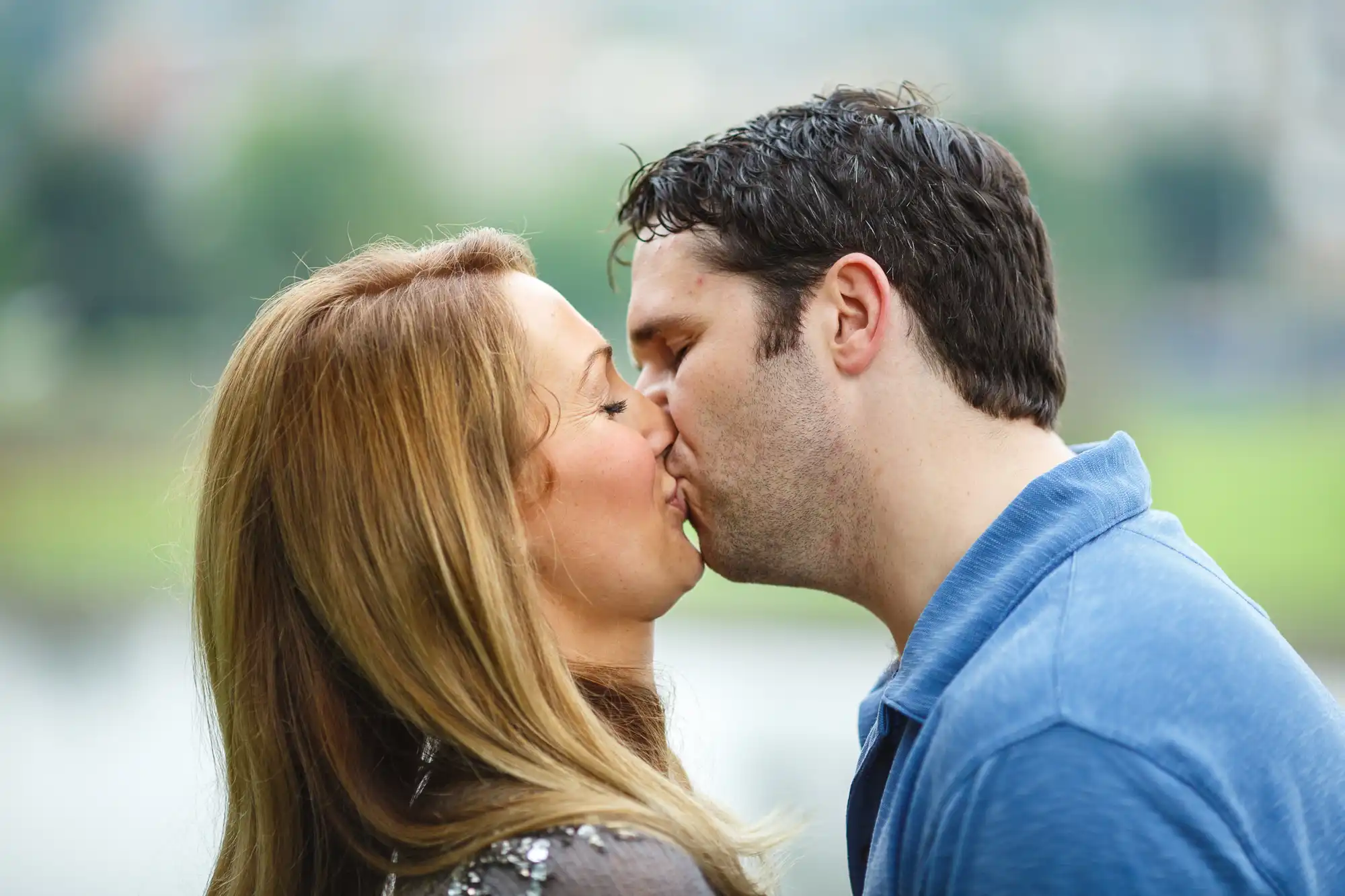 A couple kissing outdoors with a softly blurred background, both wearing casual outfits.