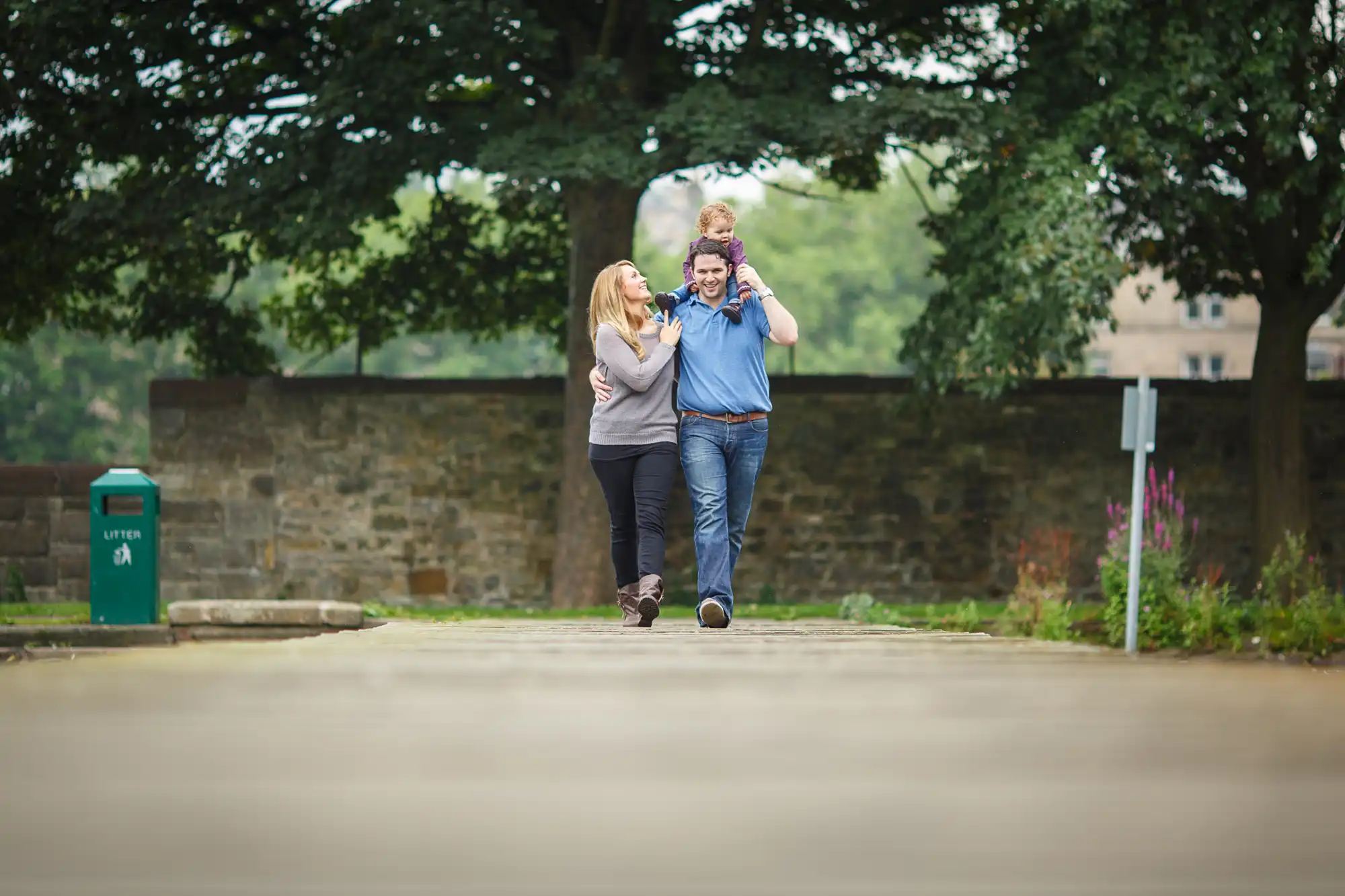A joyful couple running towards the camera on a paved path, with the woman piggybacking on the man, in a lush park setting.
