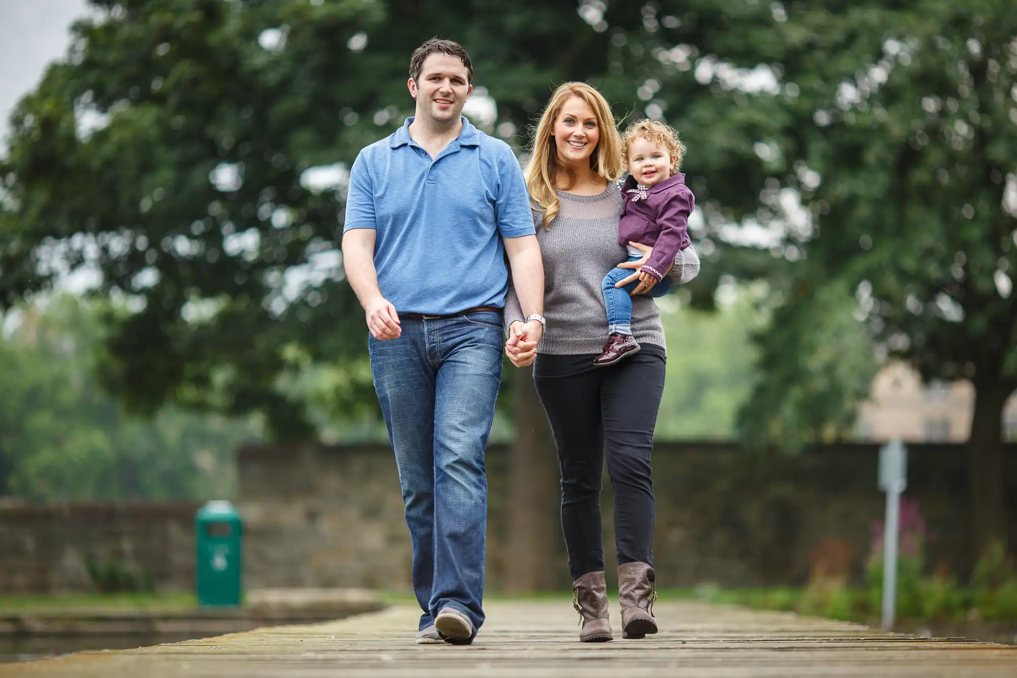 A couple walks on a bridge holding hands and carrying a toddler, surrounded by lush greenery.