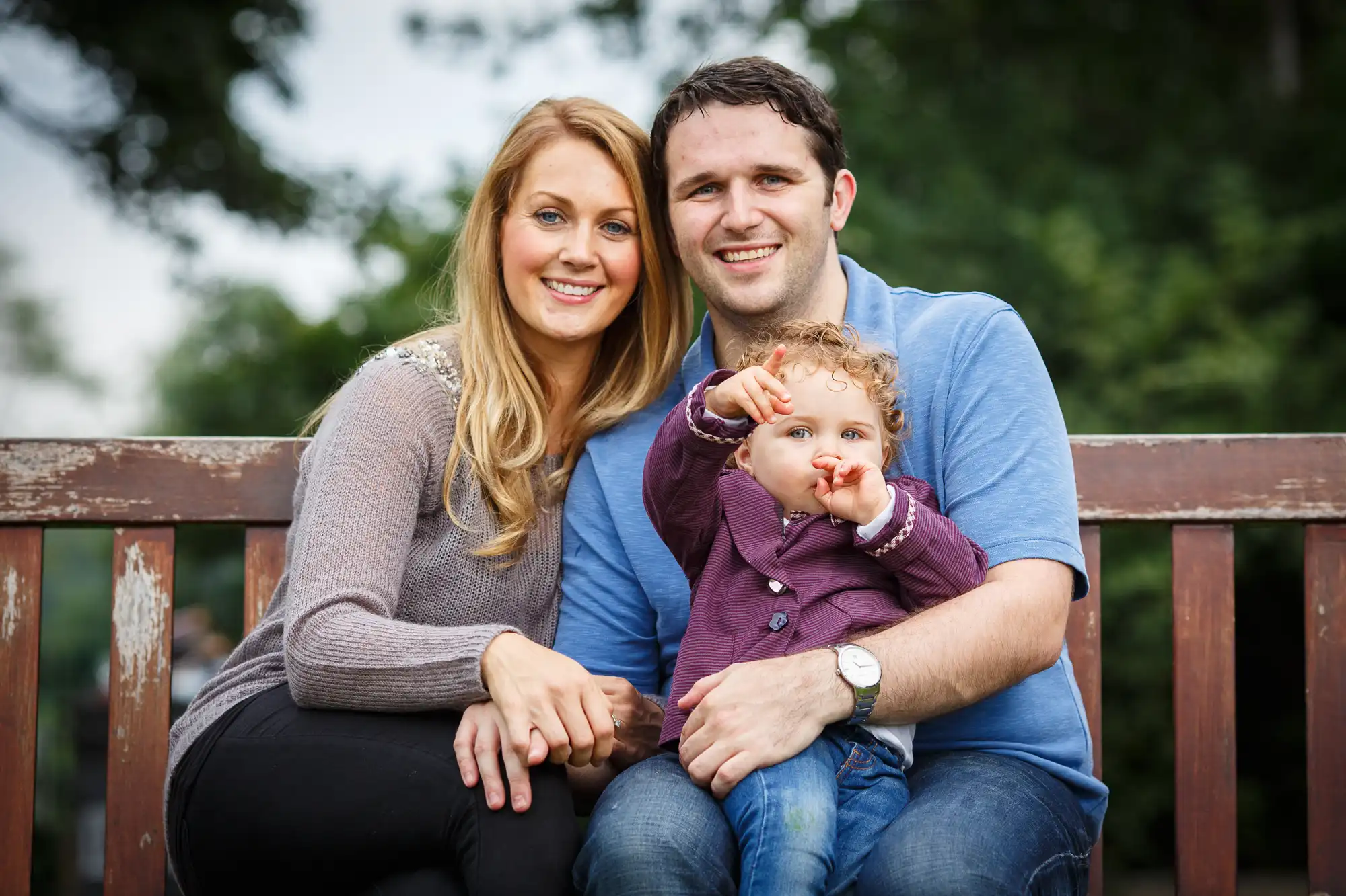 A young family with a toddler sitting together on a park bench, smiling at the camera.