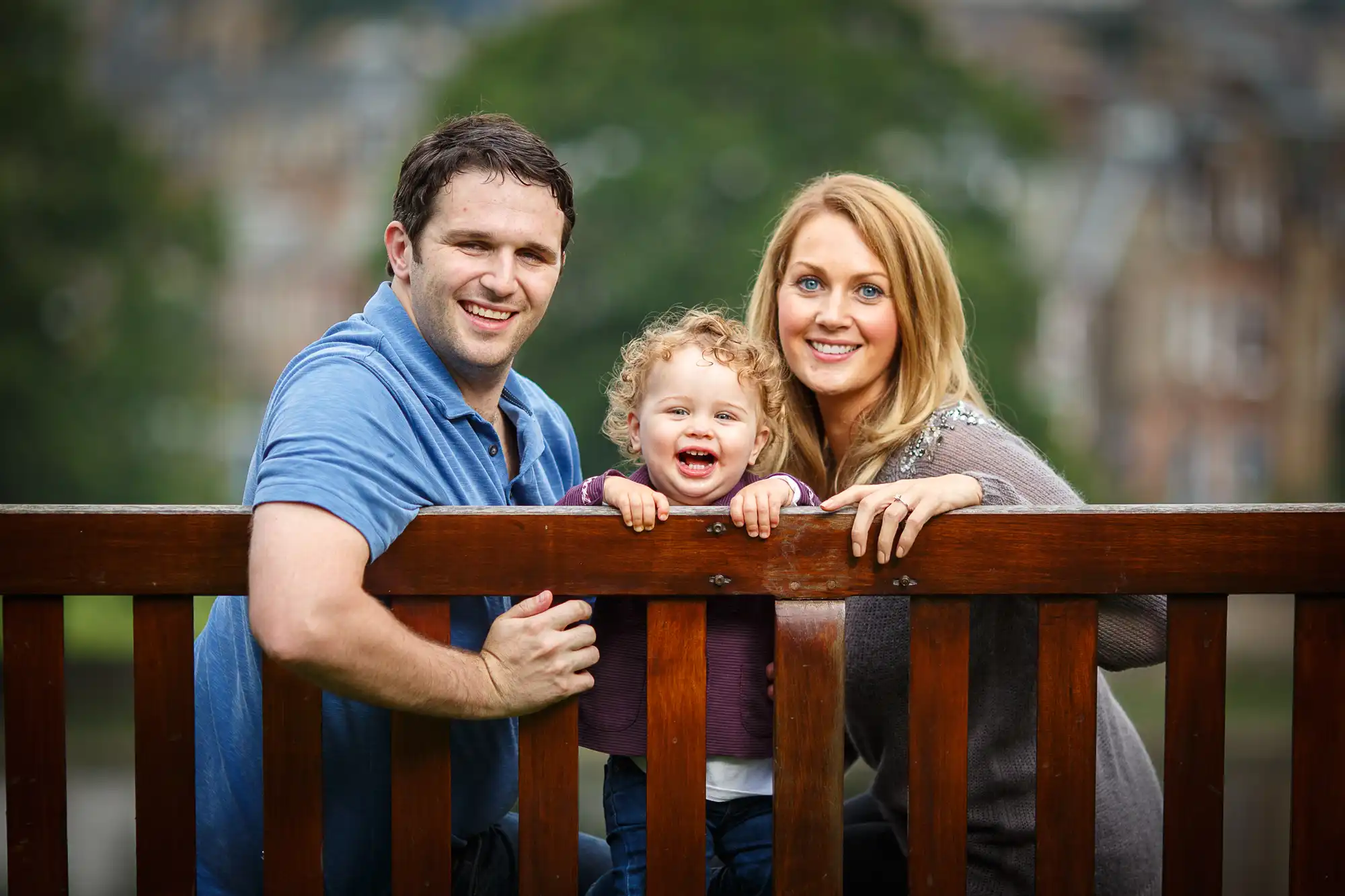 A happy family with a child standing behind a wooden railing, smiling outdoors with a blurred background of trees and buildings.