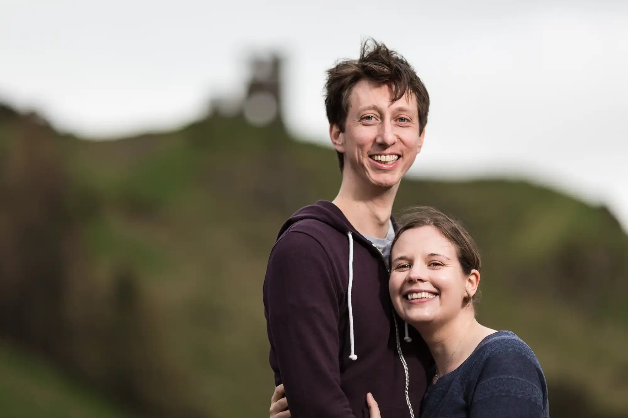 A smiling young couple embracing outdoors with a blurred historic tower and green hills in the background.