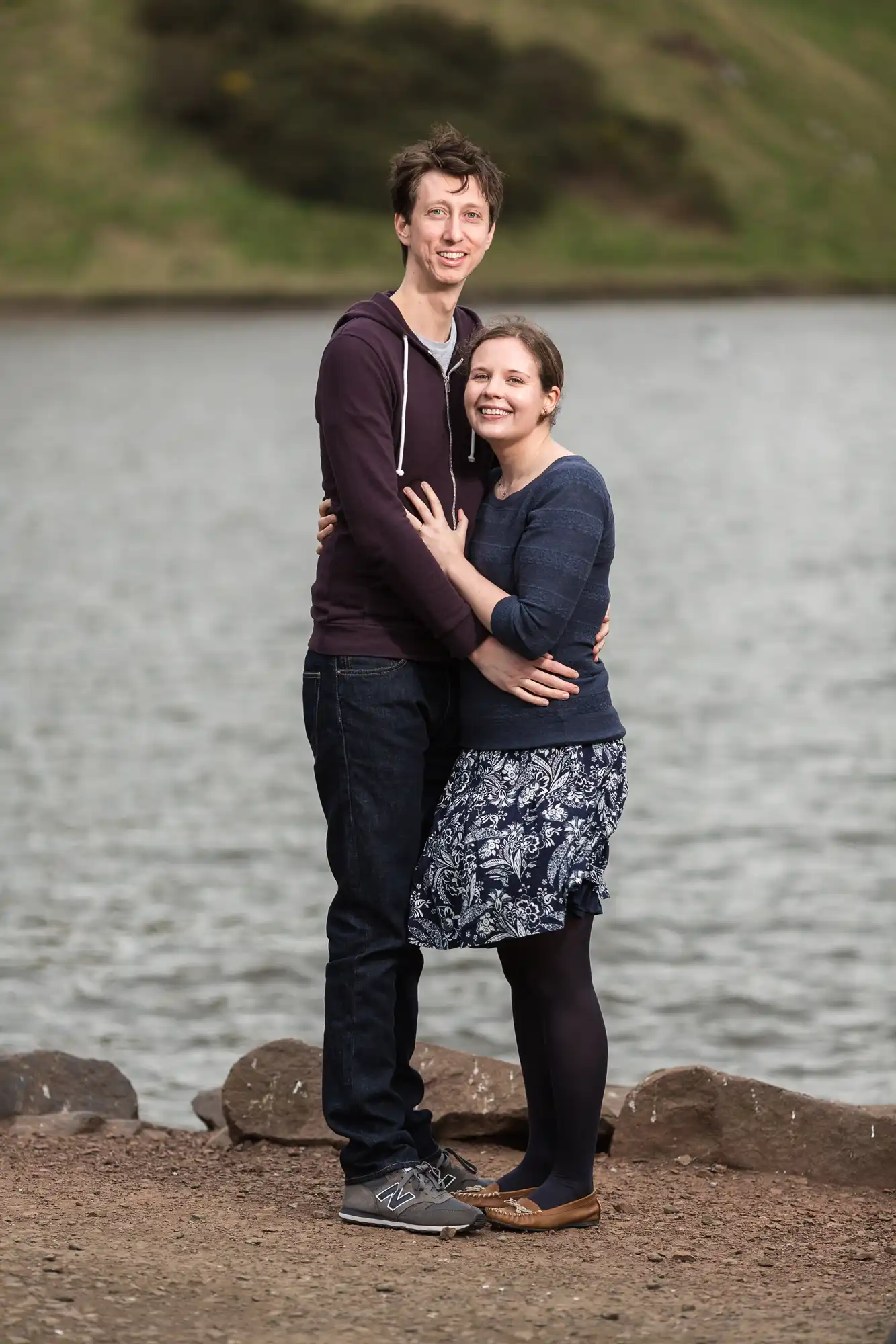 A couple embracing and smiling by a lakeside, with a hill in the background. the man is wearing a purple hoodie and jeans, and the woman a blue top with a floral skirt.