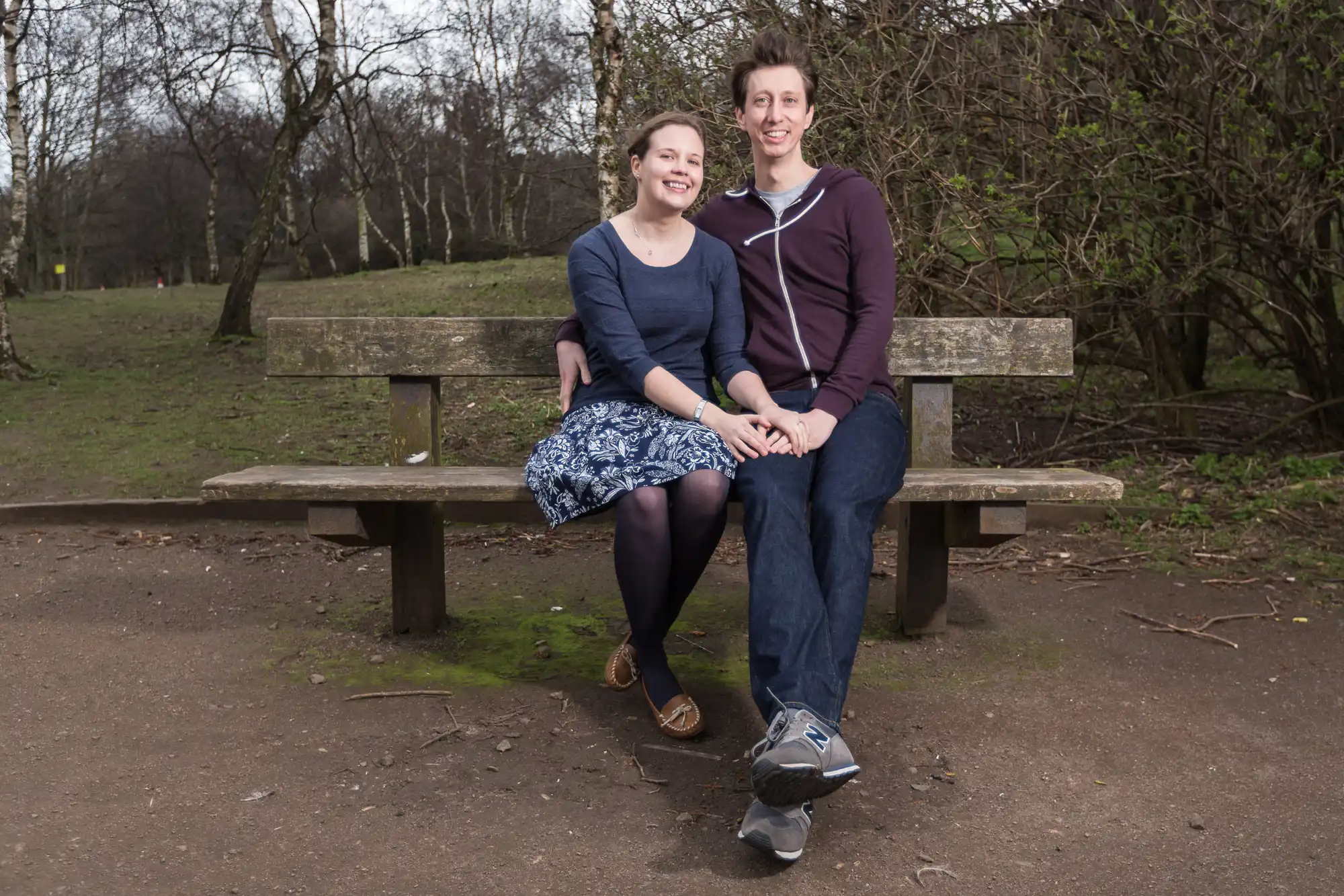 A man and a woman sitting closely on a park bench, smiling at the camera. both are dressed casually, surrounded by trees with some bare branches.