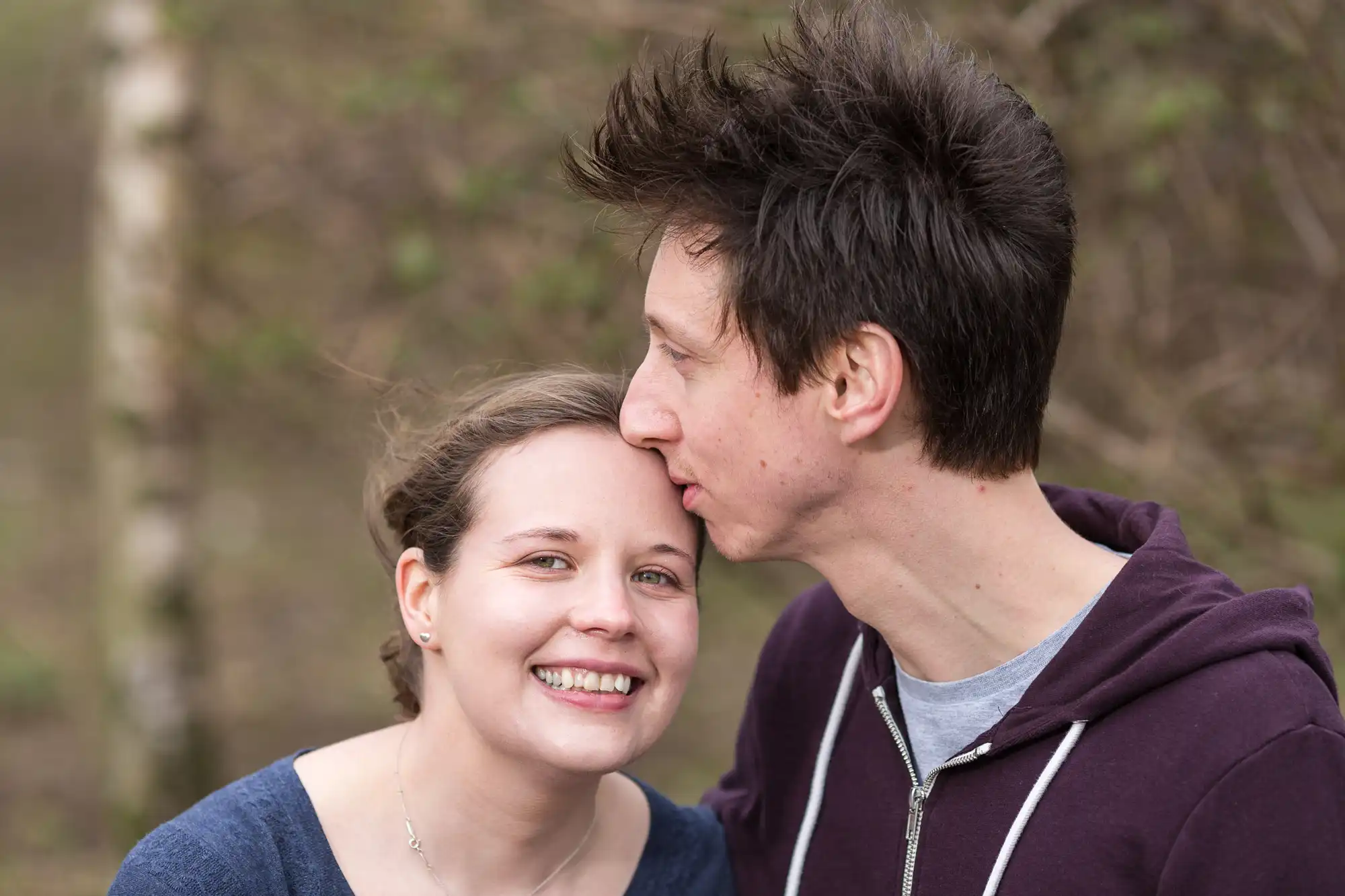 A man kissing a smiling woman on the cheek outdoors, both wearing casual clothing with a blurred natural background.