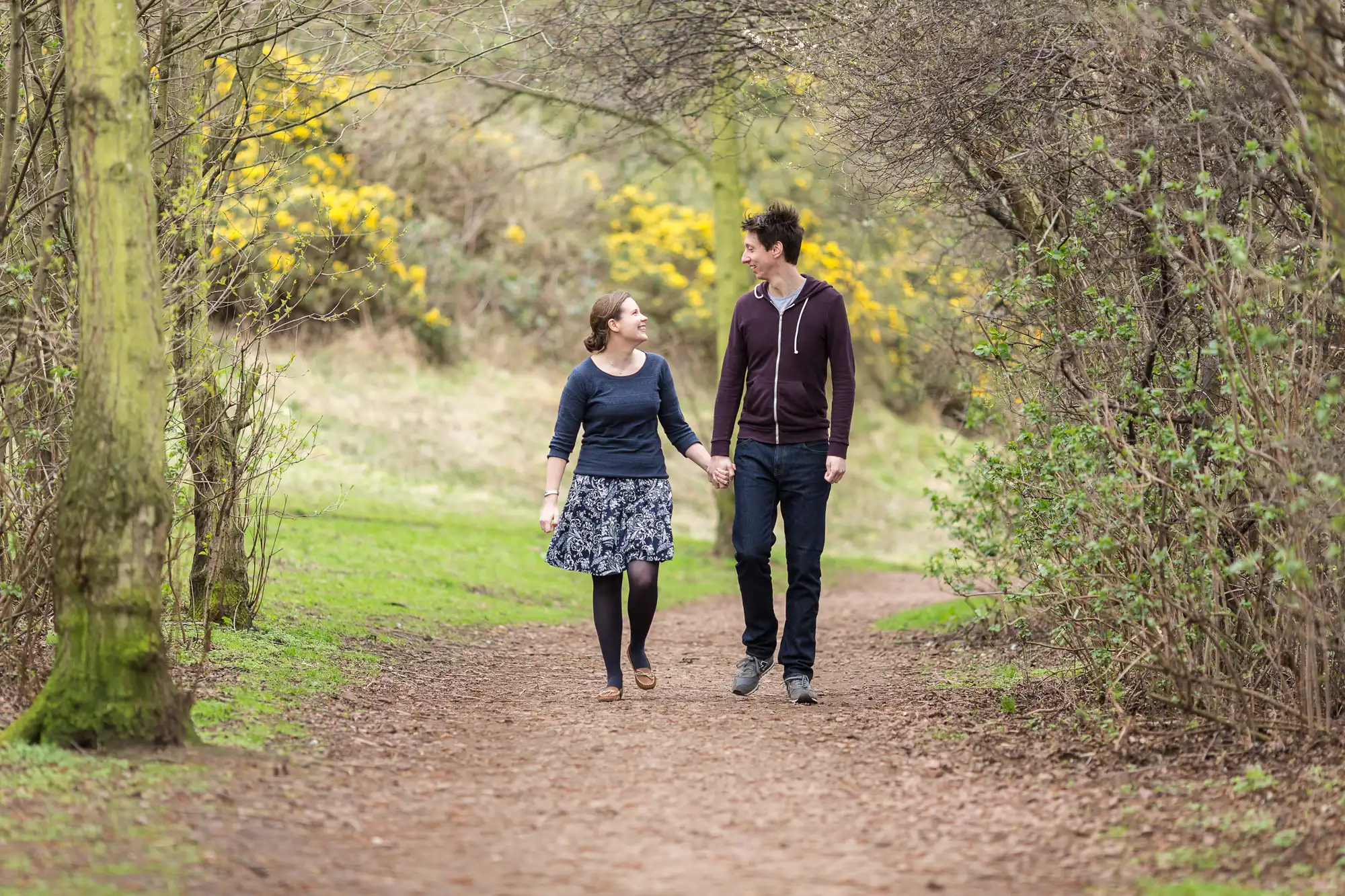 A couple holding hands while walking on a forest path, with yellow flowers in the background.