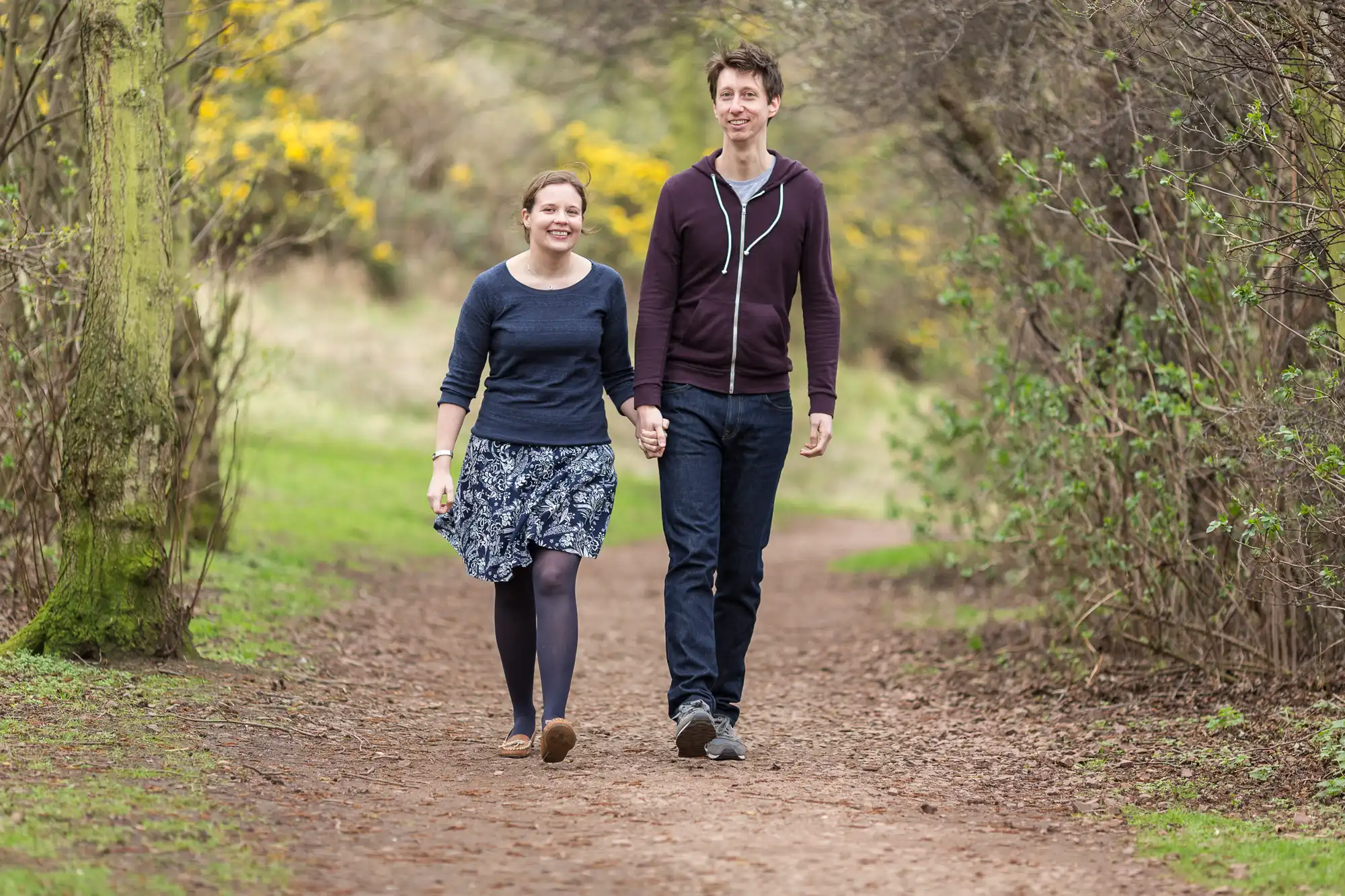 A young couple holding hands and smiling while walking on a dirt path in a lush park.