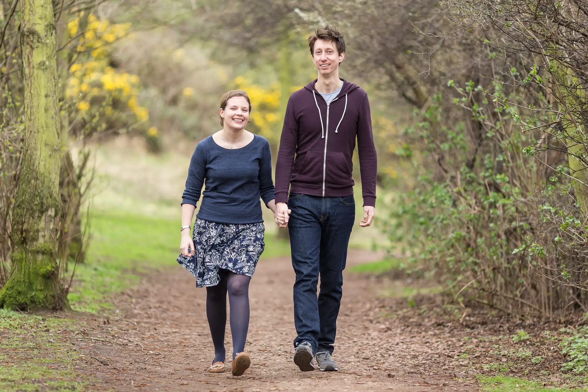 A young couple walking hand in hand down a dirt path surrounded by greenery and blooming yellow shrubs.