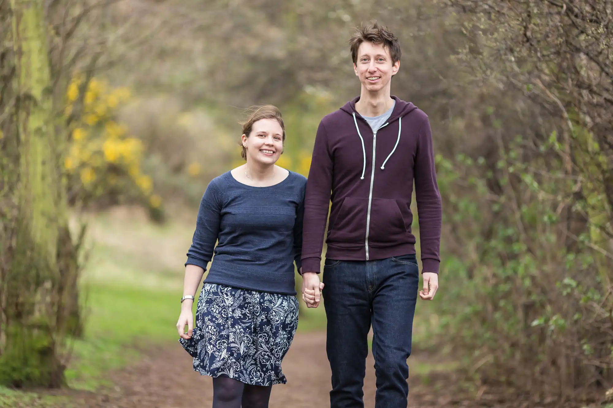 A young couple holding hands and smiling while walking on a forest path, surrounded by trees and undergrowth.