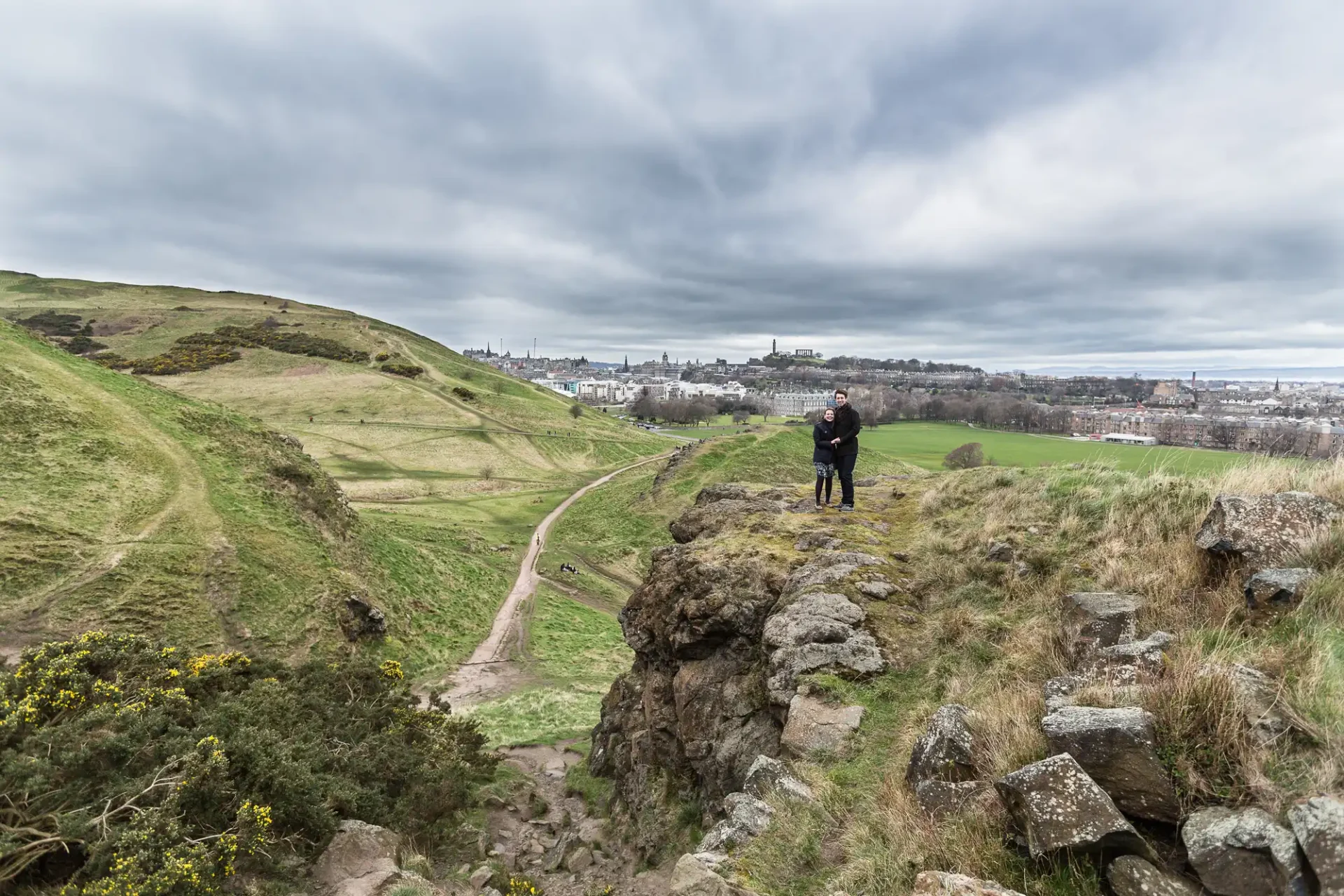 pre-wedding photoshoot at Arthur's Seat - Jon and Mandy: Two people stand on a rocky outcrop overlooking a vast grassy landscape with a distant city skyline under a cloudy sky.