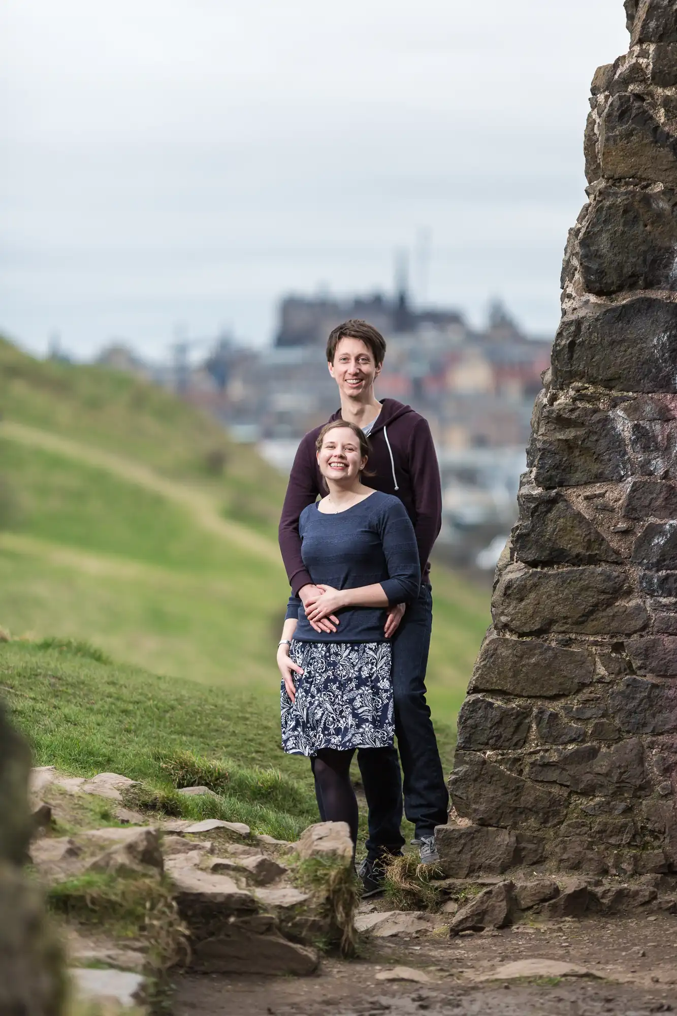 A couple smiling and posing together on a grassy hill, with a historic cityscape blurred in the background.
