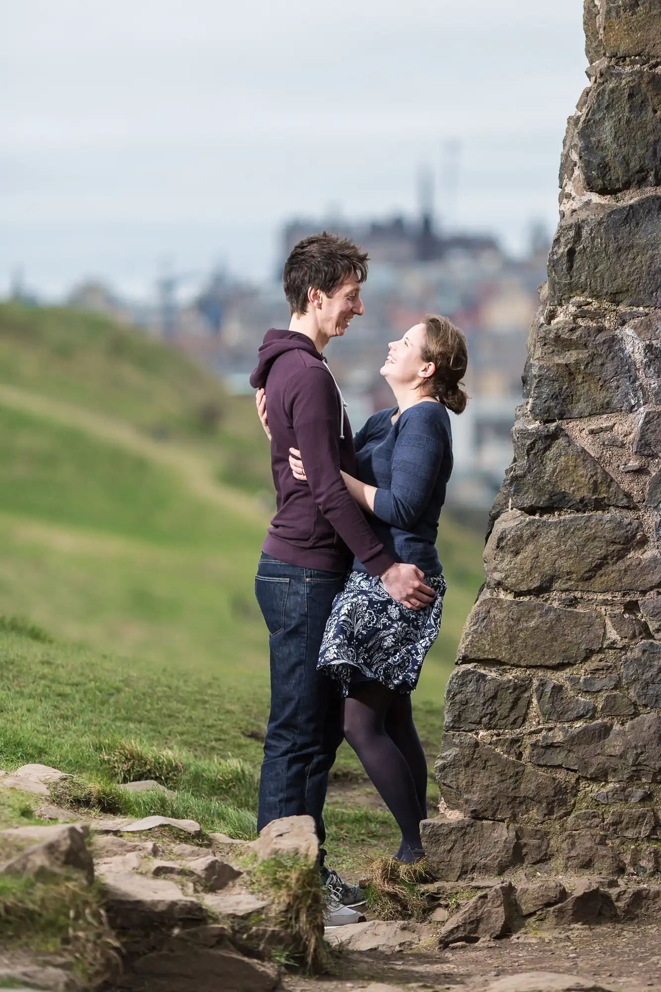 A couple embracing and smiling at each other next to a stone wall on a grassy hill, with a cityscape in the distant background.