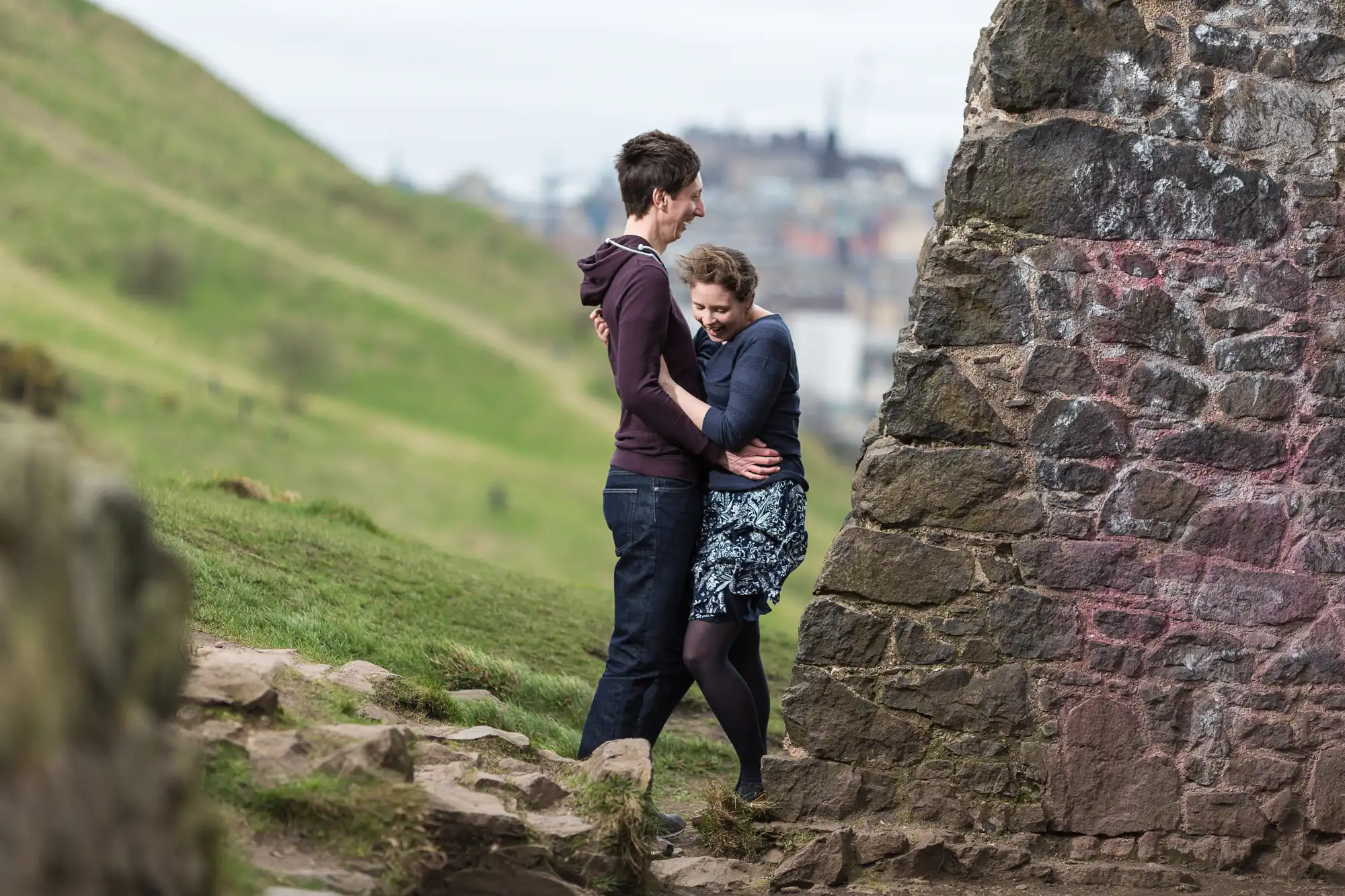 A couple embracing near a graffiti-covered stone wall, with a grassy hill and distant cityscape in the background.