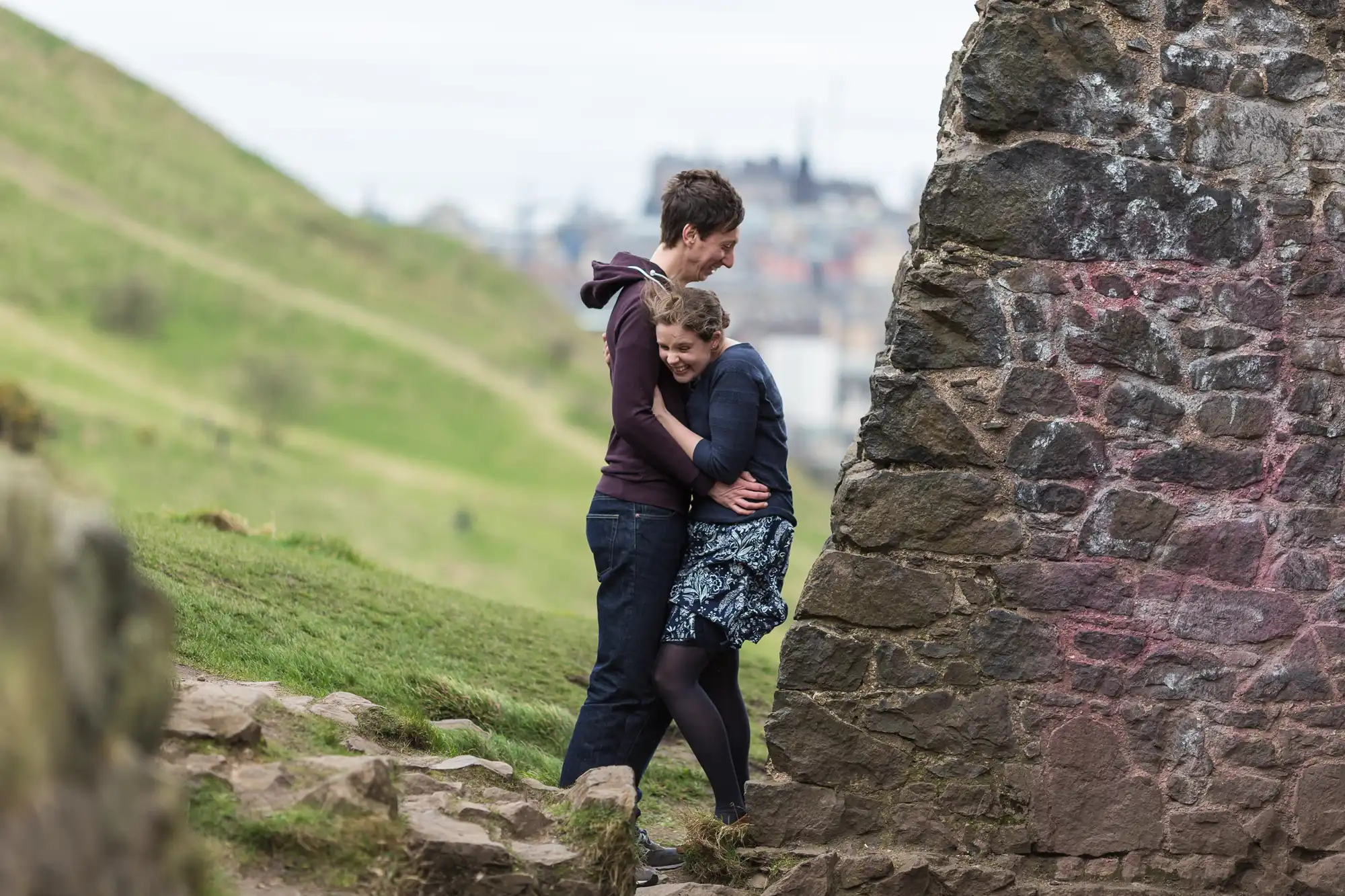A couple embracing near a graffiti-covered stone wall with a grassy hill and cityscape in the background.