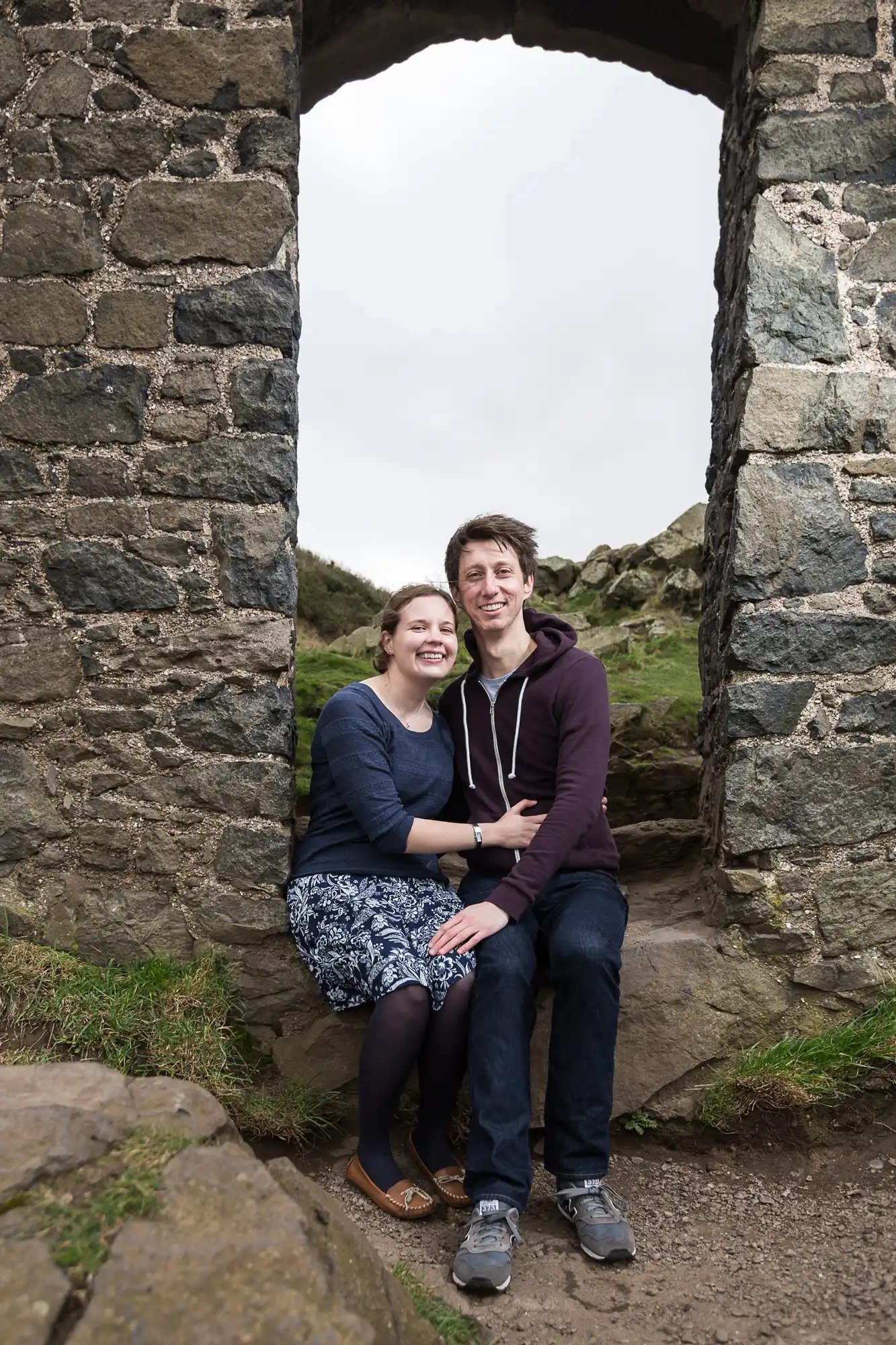 A couple smiling and sitting together in an archway of a stone ruin, surrounded by grassy hills.
