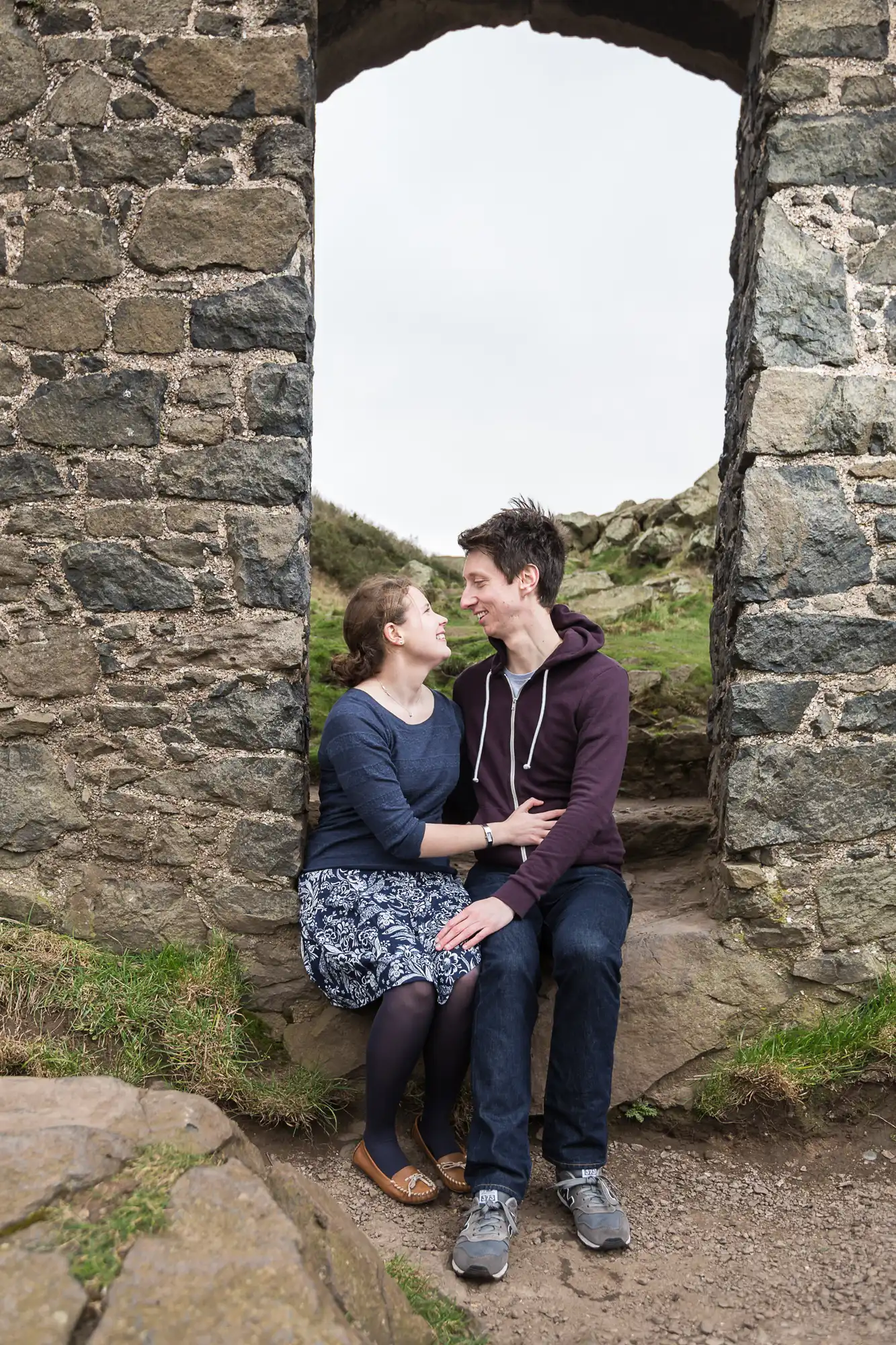 A couple sitting in a stone archway lovingly looking at each other, with a grassy hill in the background.