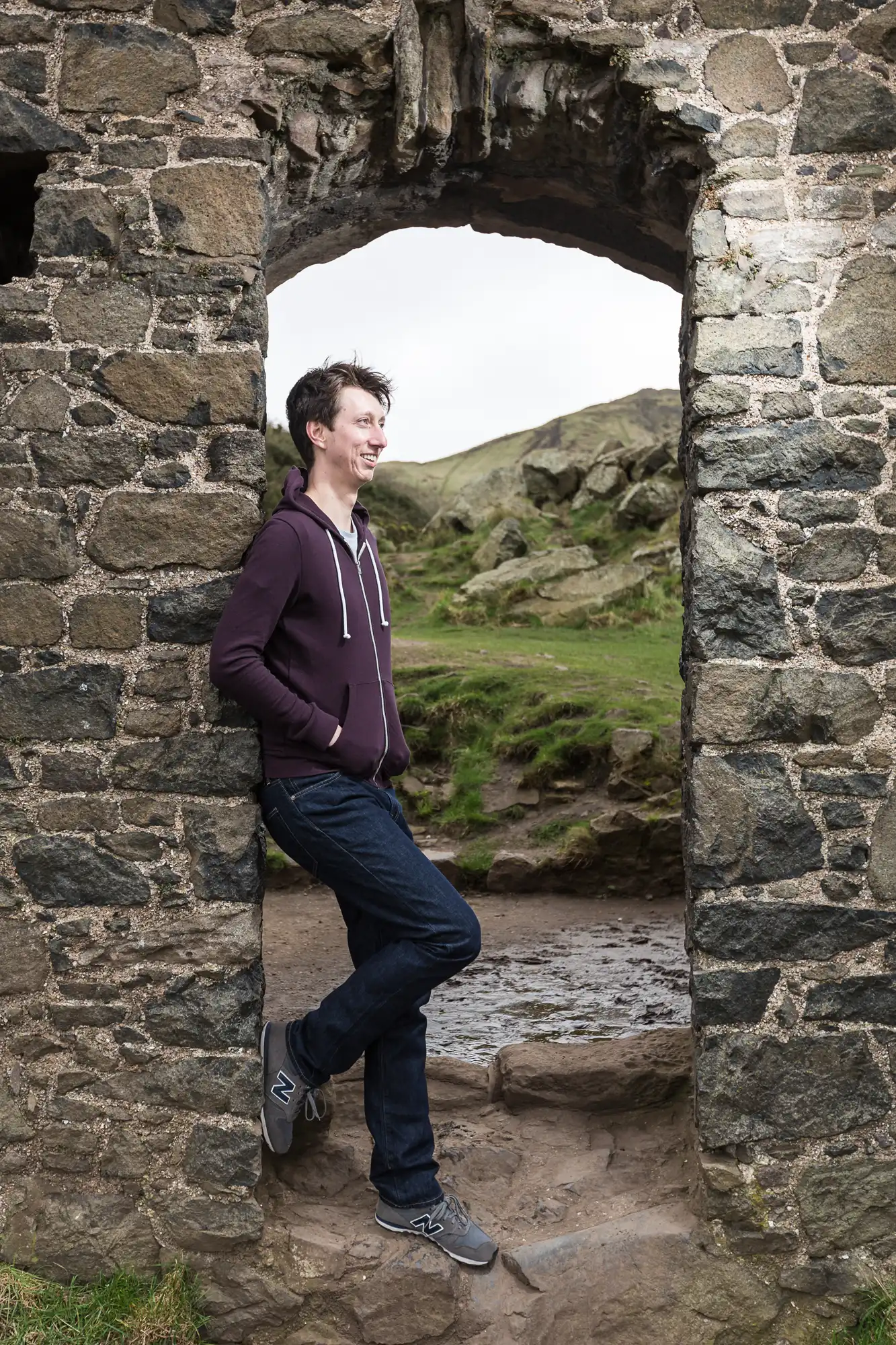 A man in a purple hoodie leans casually in an arched stone doorway, looking out onto a grassy landscape.