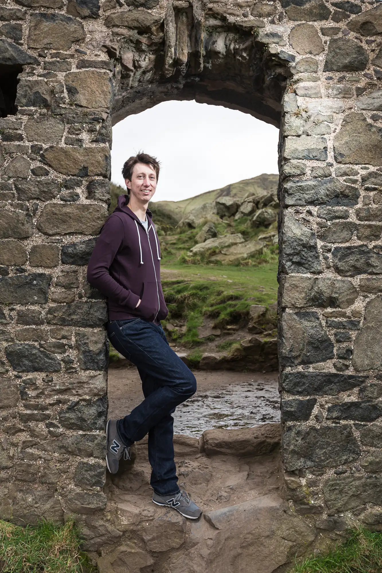 A man leaning in a stone archway with a natural landscape in the background. he is smiling, dressed casually, and has one foot elevated on the arch.