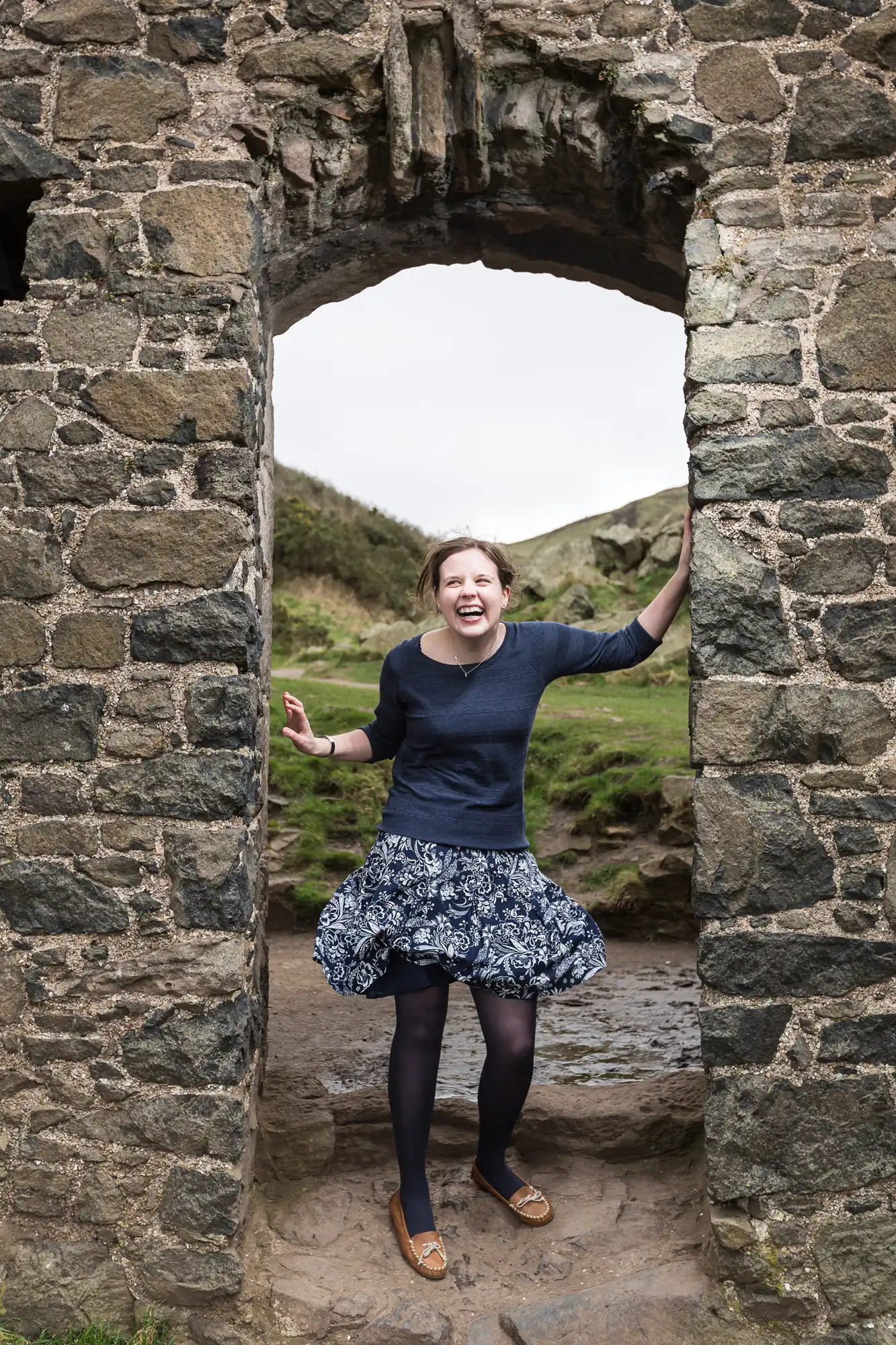 A joyful woman in a dress and tights stands in an open stone doorway, laughing with arms extended, amid a grassy landscape.