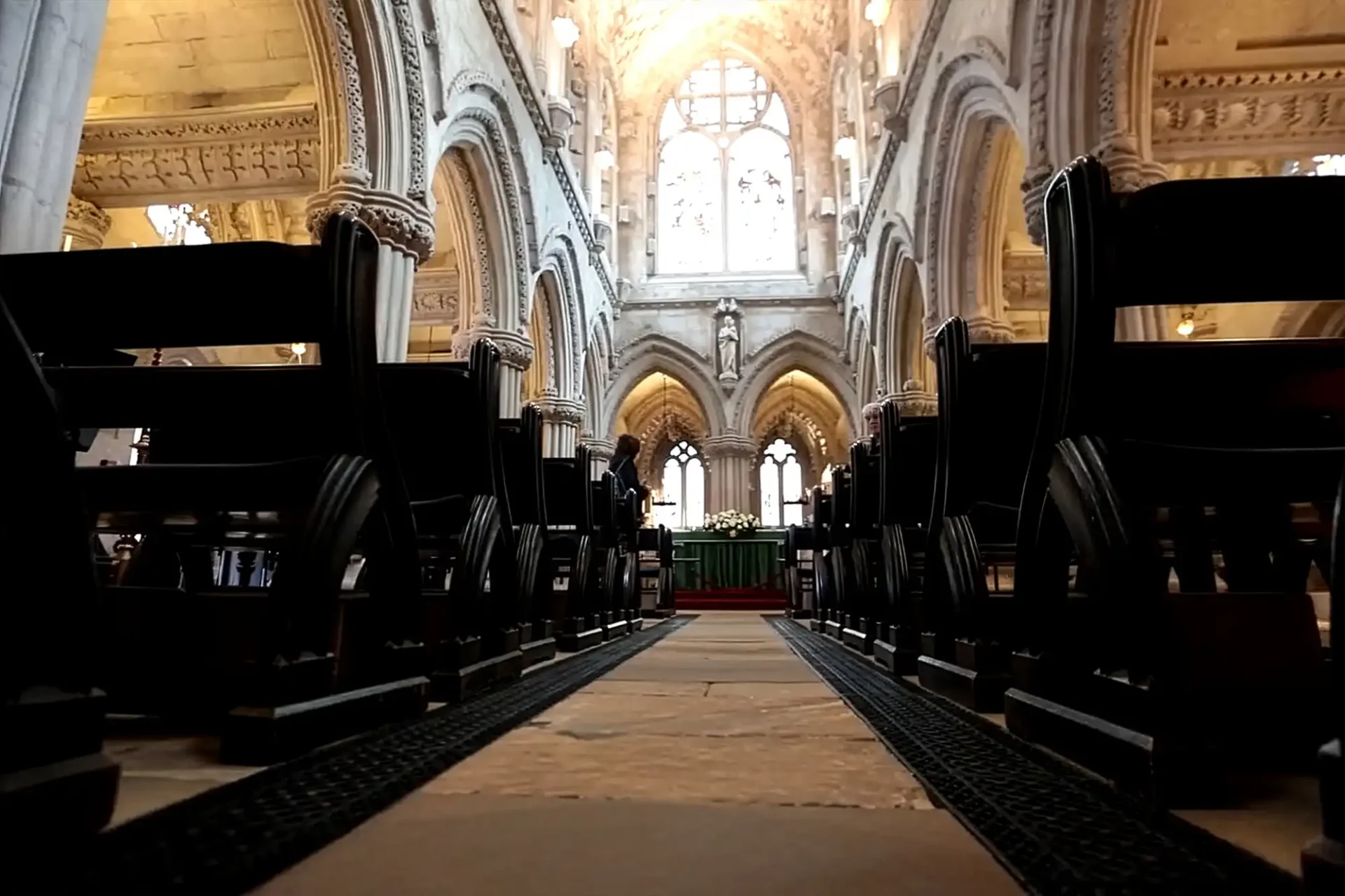 Low-angle view of a grand Rosslyn Chapel interior, showing ornate stone arches, tall columns, and stained glass windows. Rows of dark wooden pews line both sides of a central aisle leading to an altar under an illuminated vaulted ceiling.