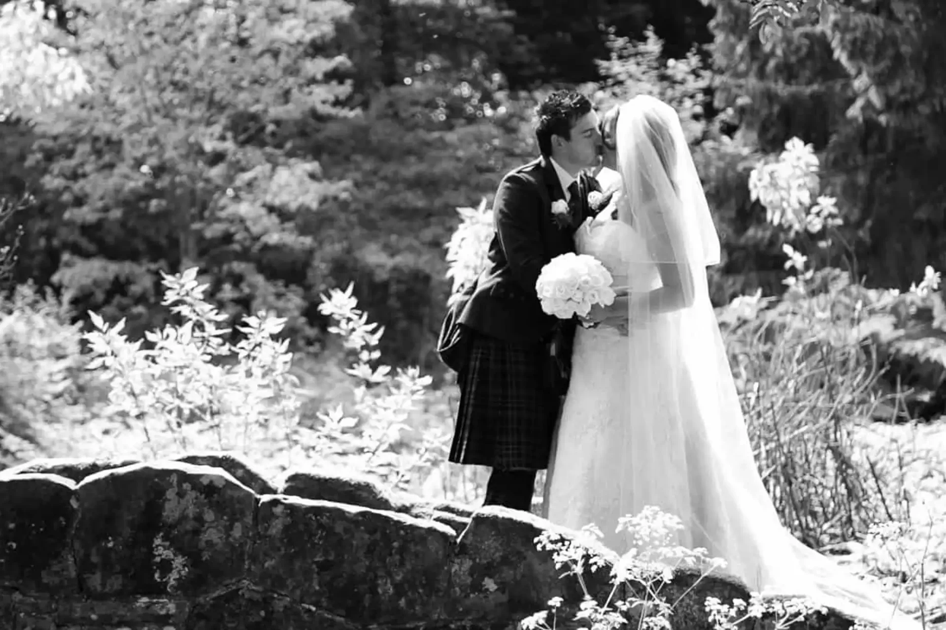 A bride in a white dress and veil holds a bouquet of flowers while kissing a groom in a dark suit and kilt. They stand on a stone bridge surrounded by leafy trees and foliage on a sunny day at Rufflets St Andrews. The image is in black and white.