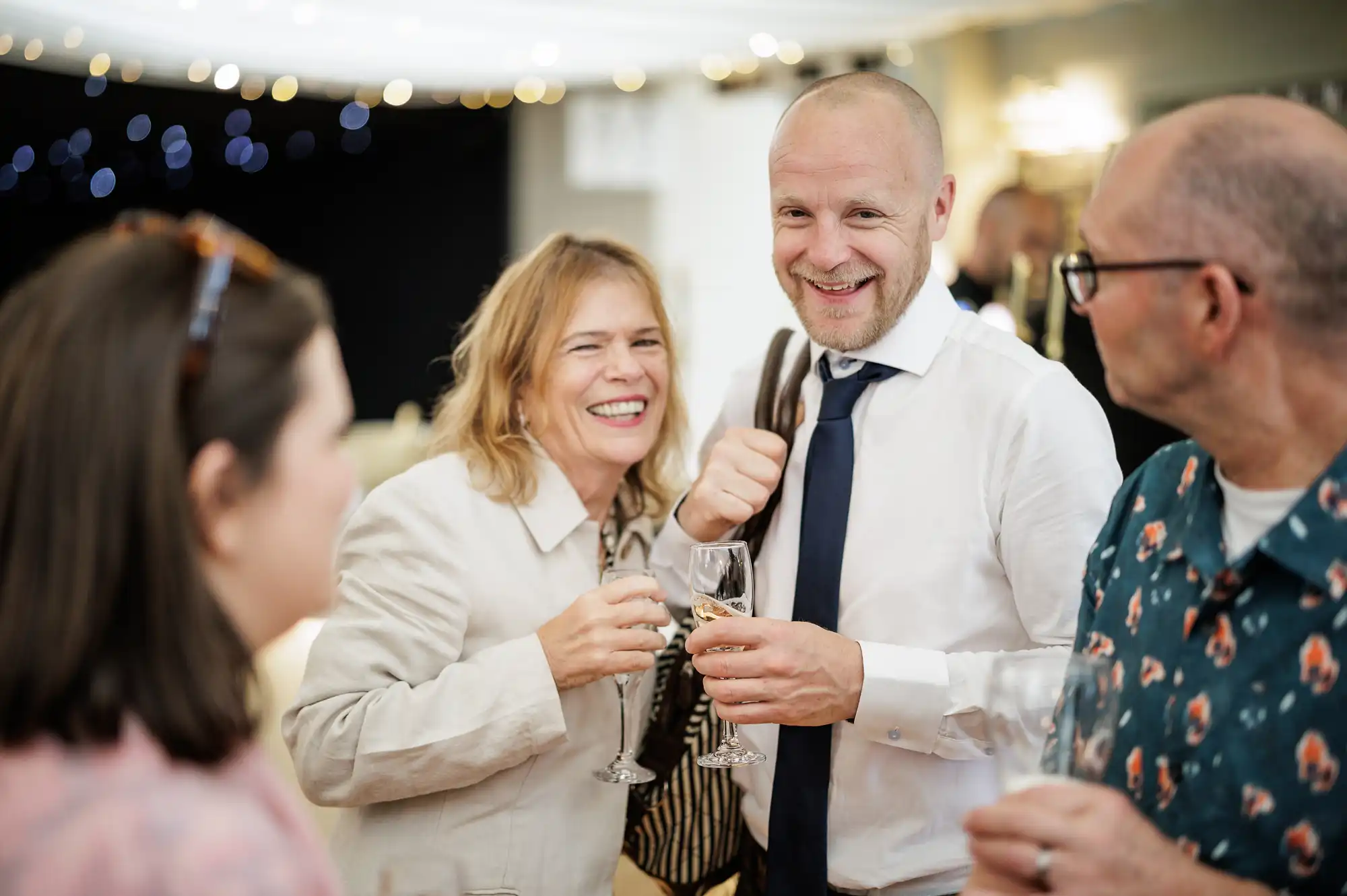 Four adults stand close together at a social event, smiling and talking whilst holding drinks. One man in a white shirt and tie gestures enthusiastically. The background is softly lit with fairy lights, creating a warm and festive atmosphere.