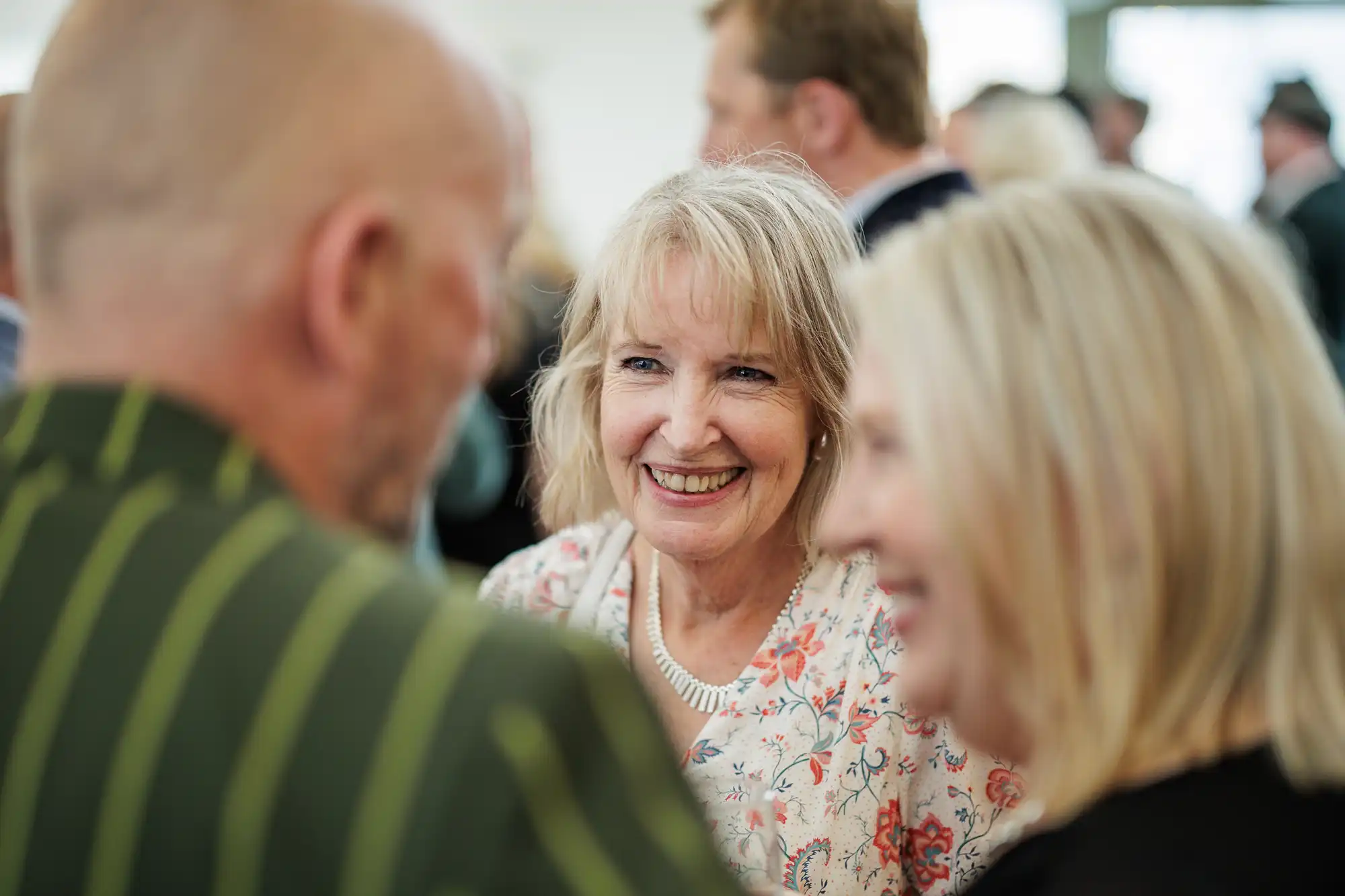 A smiling older woman with fair hair, wearing a floral top, stands indoors among a group of people. She looks at a bald man in a striped jacket and a blonde woman, both slightly out of focus in the foreground, suggesting a friendly conversation.