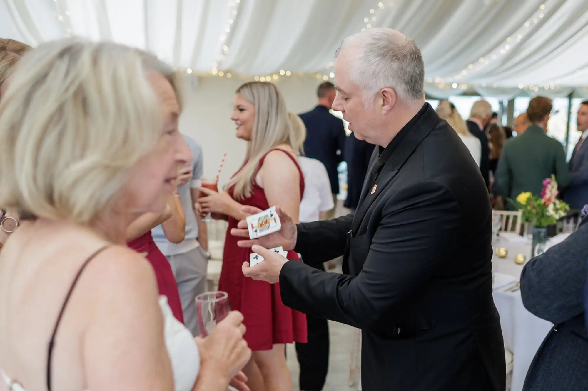 A magician in a black suit performs a card trick for a group of guests at a formal event under a white draped marquee with fairy lights. A woman in a white dress holds a drink and watches closely. Other guests chat and mingle in the background.
