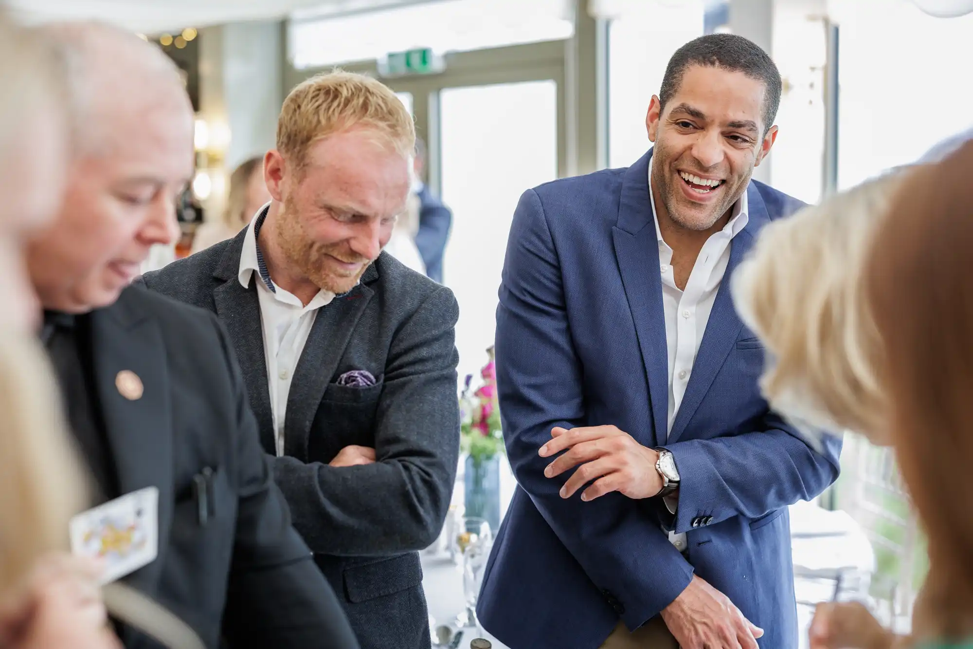 Three men, dressed in business attire, stand together at a bright indoor event. One man in a blue blazer smiles widely, another with a beard and a grey suit laughs, and a third man in black looks down. The setting is lively, with blurred people and flowers in the background.