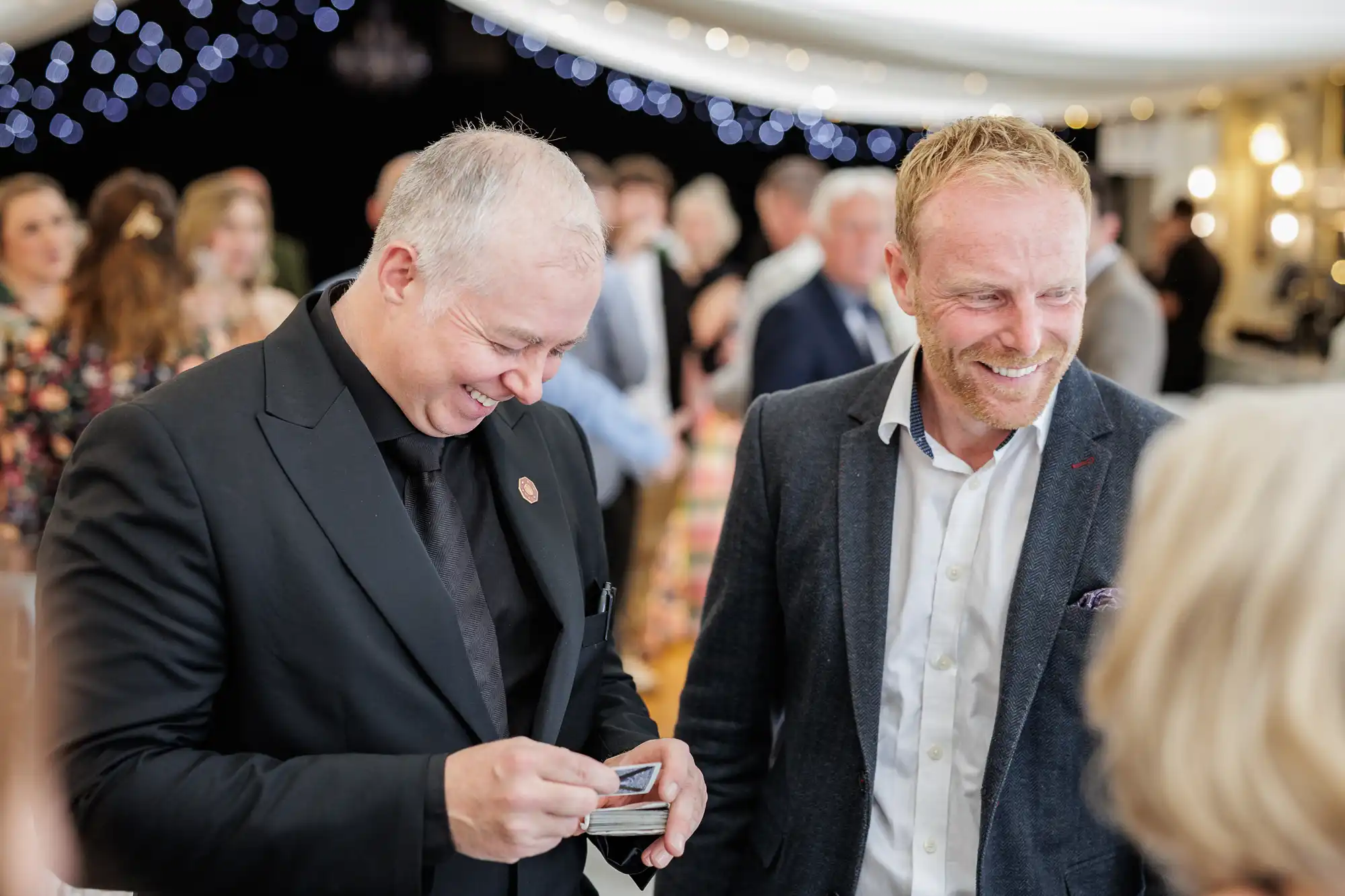 Two men in suits smile and laugh at a lively gathering. One man, holding playing cards, appears to perform a magic trick. Blurred guests and sparkling fairy lights fill the festive background, creating a warm, joyful atmosphere.