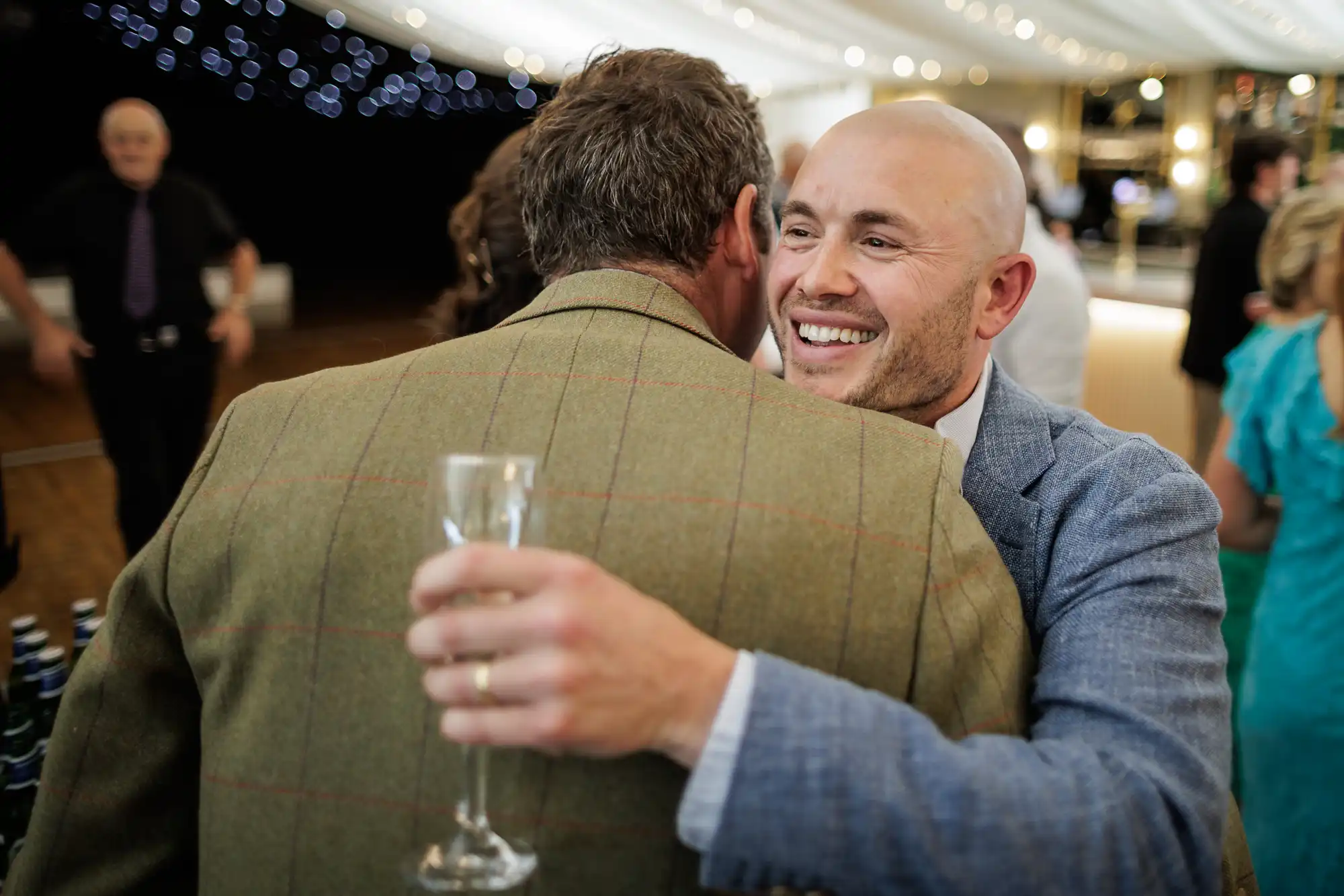 A smiling bald man in a light suit embraces another man in a brown checked jacket at a lively indoor event, holding a champagne flute. Fairy lights and blurred guests in formal attire create a festive atmosphere in the background.