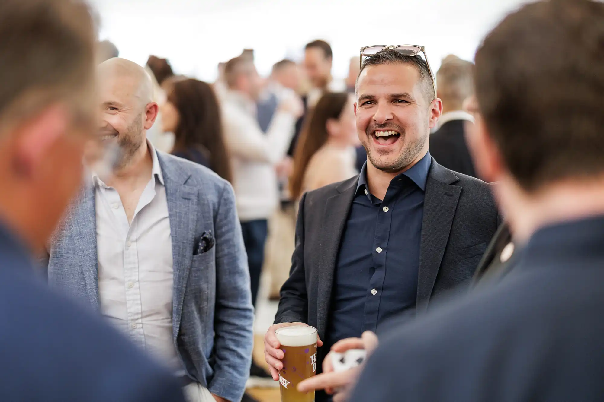 A man in a dark suit smiles and laughs whilst holding a drink at a crowded indoor gathering. Other people, mostly dressed in smart-casual attire, talk and mingle in the blurred background. The atmosphere appears lively and sociable.
