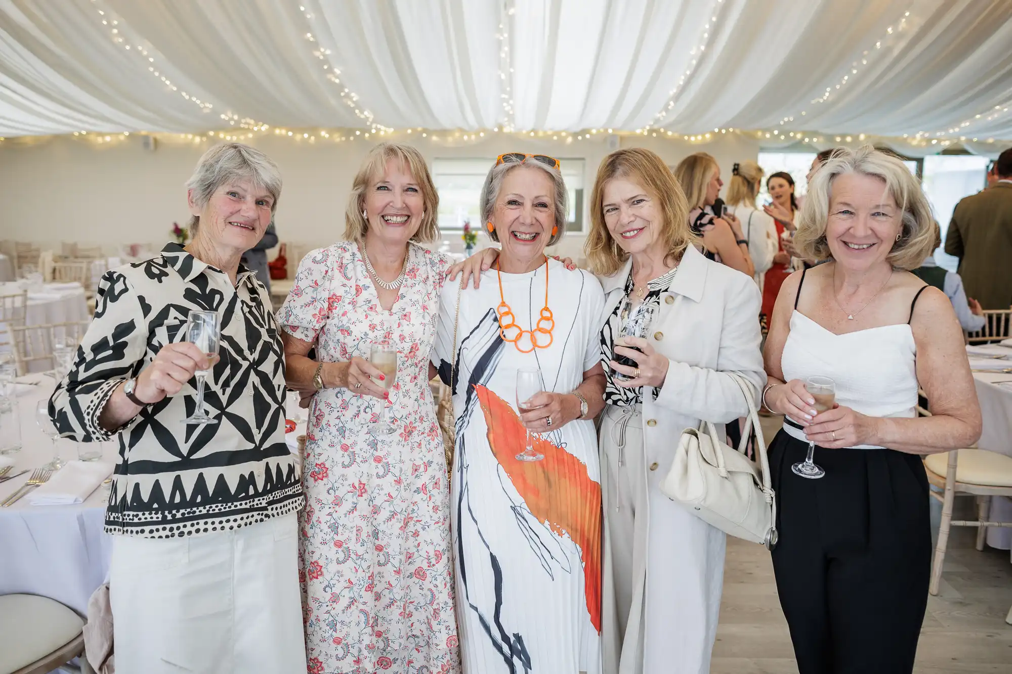 Five smiling women pose together at an indoor event, holding drinks. They wear elegant, light-coloured outfits. The venue is decorated with white drapes and strings of lights, and round tables with white linen are visible in the background.
