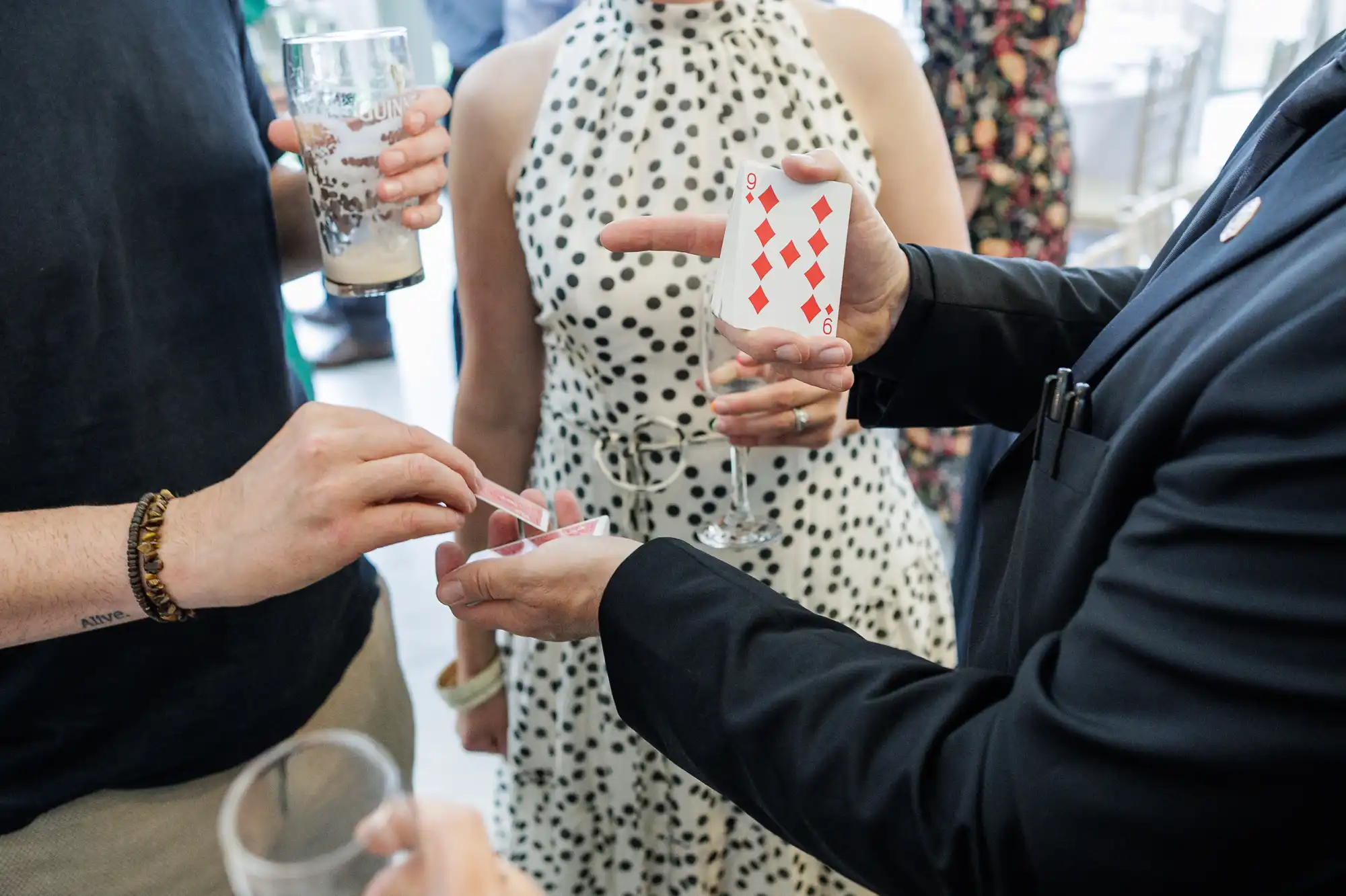 A magician in a suit holds up a nine of diamonds playing card while performing a trick for two people, one in a sleeveless polka-dot dress. The magician’s other hand holds a small clear box; people nearby hold drinks and appear engaged in the performance.