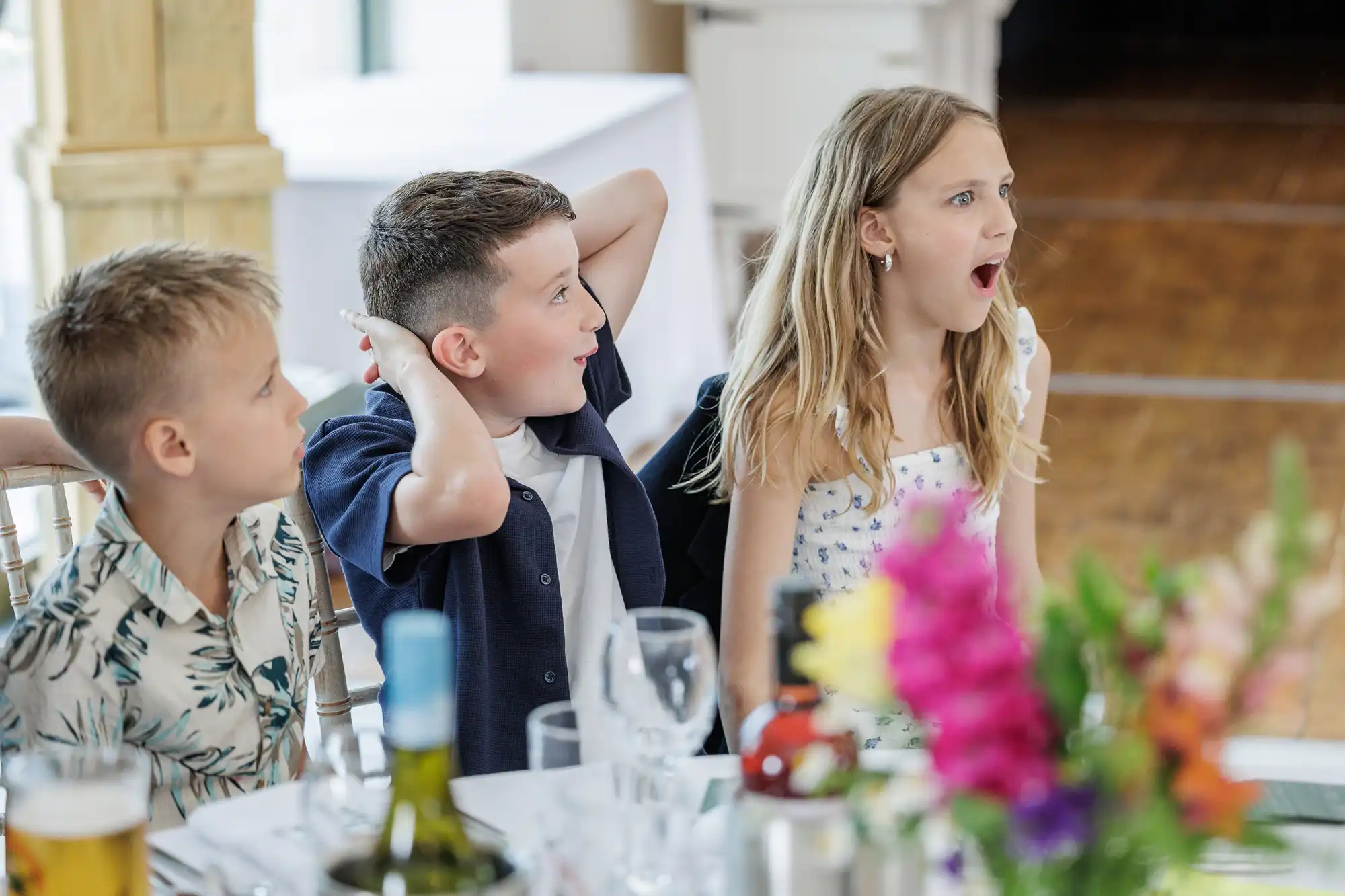 Three children sit at a table set for a meal, reacting with surprise. The boy on the left looks forward, the boy in the middle holds his hands behind his head with wide eyes, and the girl on the right has her mouth open. Bright flowers are in the foreground.