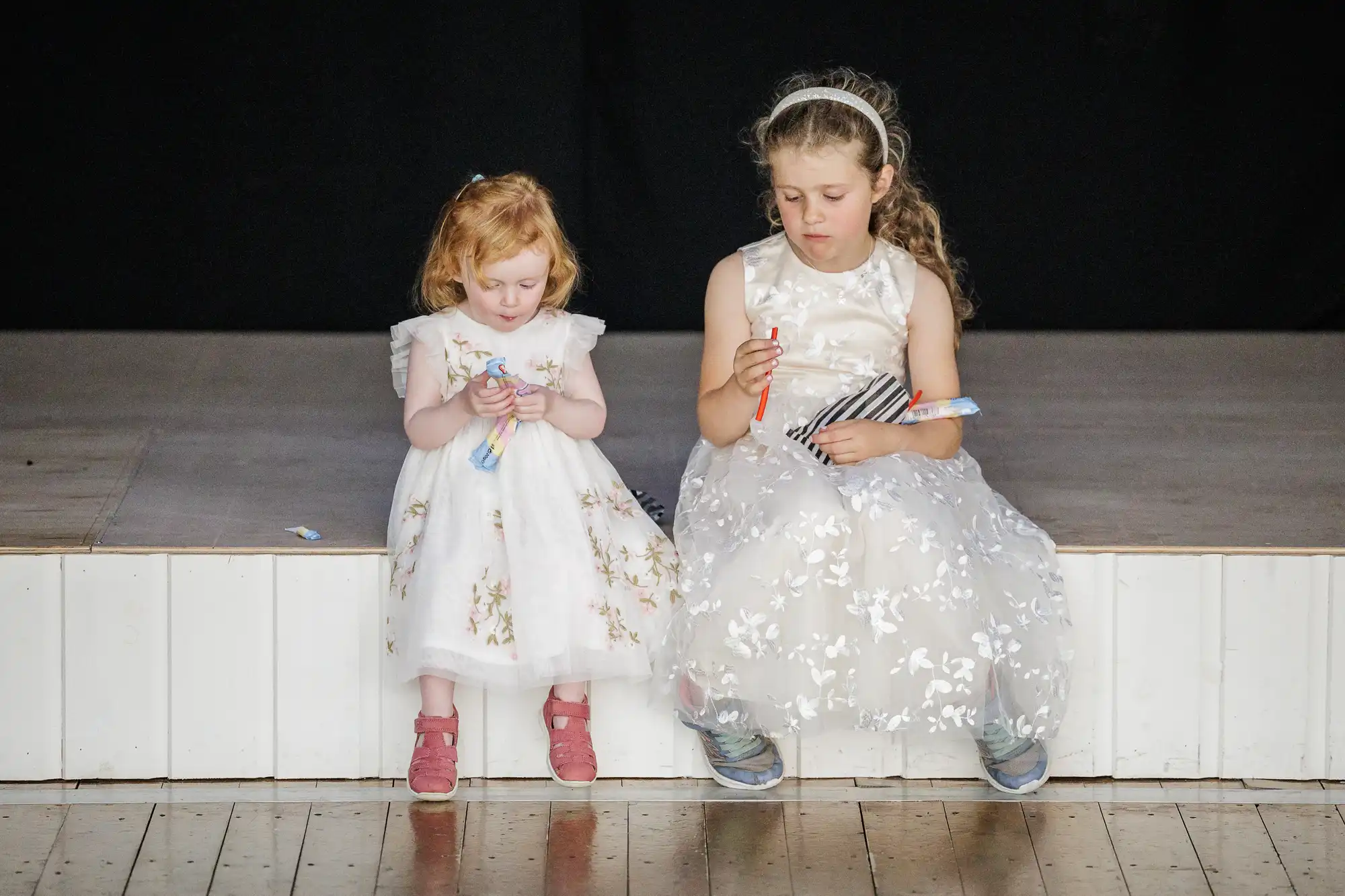 Two young girls in white, floral dresses sit on the edge of a stage. The girl on the left has red hair and pink shoes, whilst the girl on the right has curly hair, a headband, and blue trainers. Both look down, focused on items in their hands.
