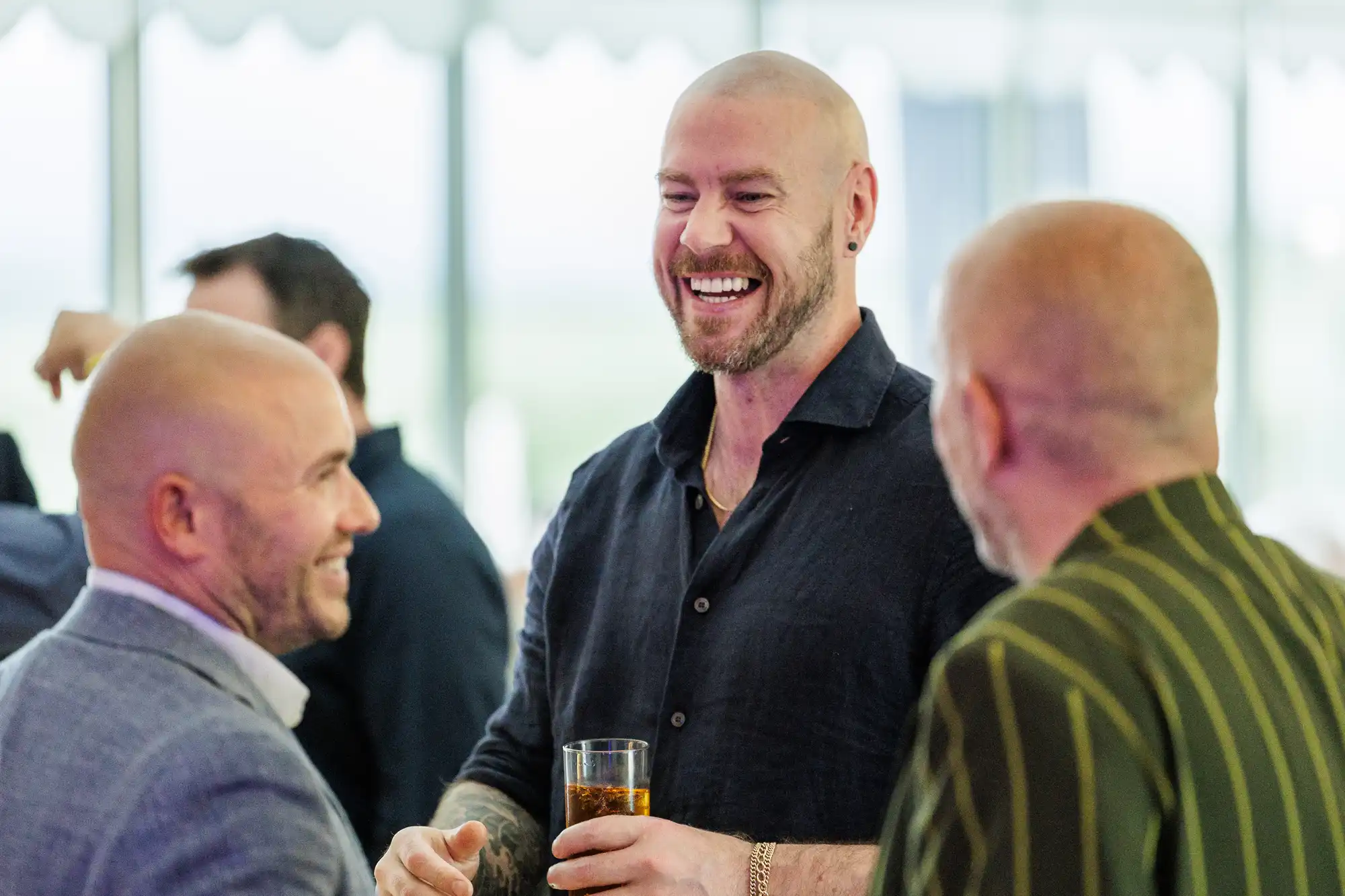 Three bald men are standing together indoors, smiling and chatting. The man in the centre, wearing a dark shirt, holds a drink and laughs. The man on the left wears a grey suit, and the man on the right wears a green striped jacket. The background is softly blurred.
