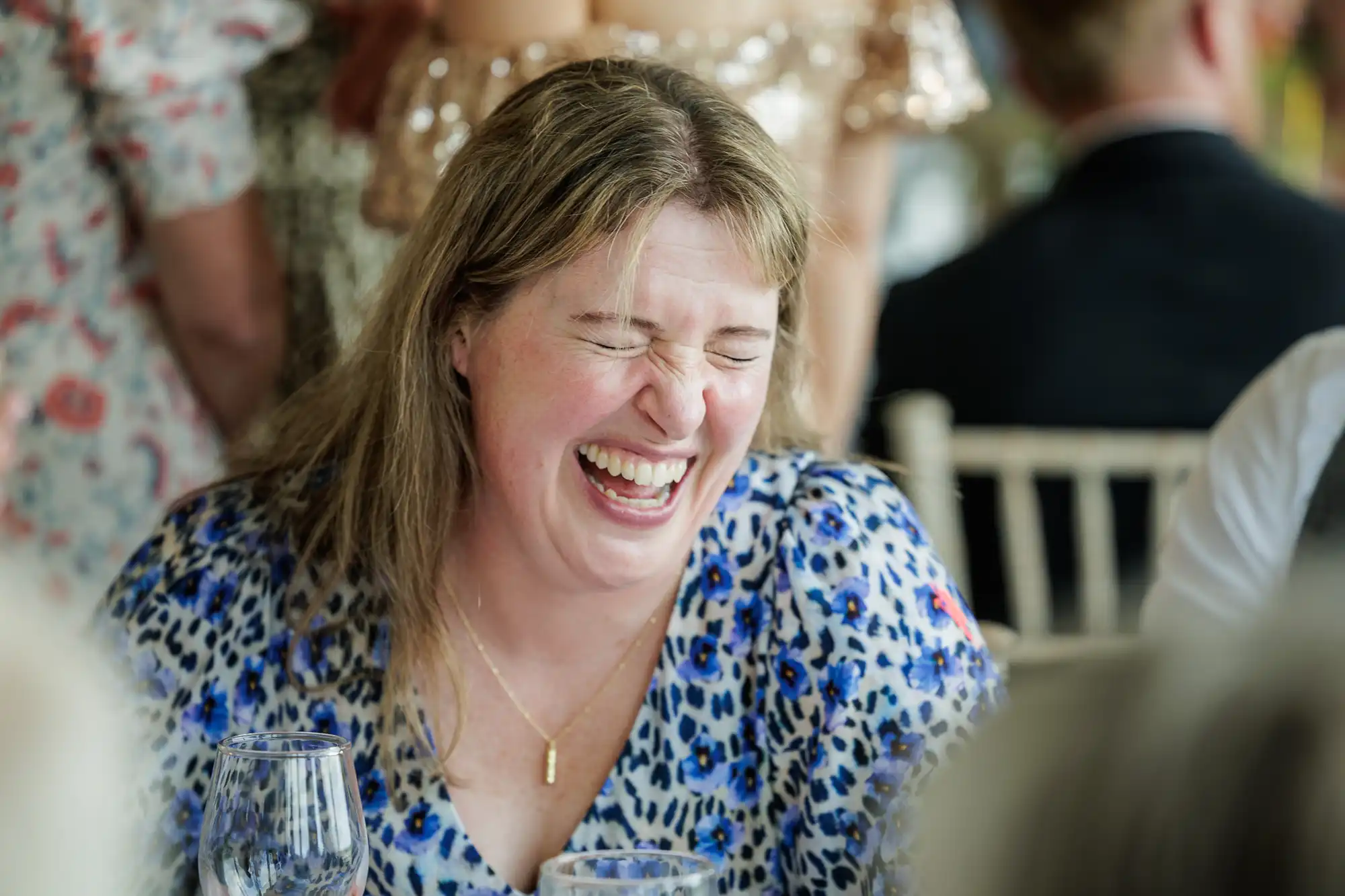 A woman with light brown hair, wearing a blue leopard print top and gold necklace, sits at a table and laughs with her eyes closed. Blurred people and chairs are in the background, suggesting a lively social gathering.
